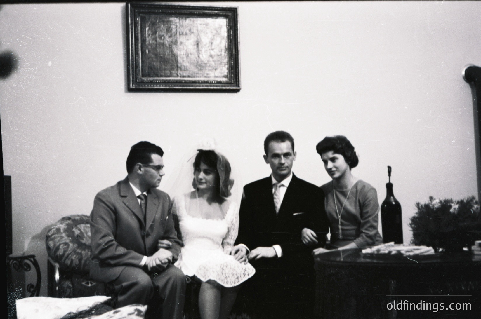 Mid-20th century wedding portrait in a formal indoor setting. Four individuals pose: bride in lace dress with veil, groom in suit, and two guests in 1950s-60s attire. Decor includes a framed artwork, vintage furniture, and a wine bottle on a side table. Classic black-and-white composition.