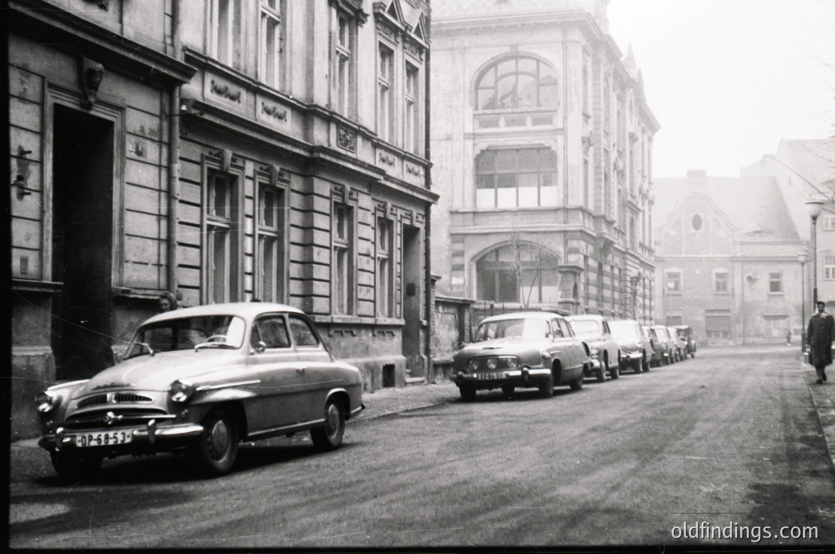 Classic 1950s European street scene with vintage cars parked along a cobblestone road. Architectural details include ornate facades with arched windows and decorative moldings. Foggy atmosphere enhances nostalgic urban vibe.