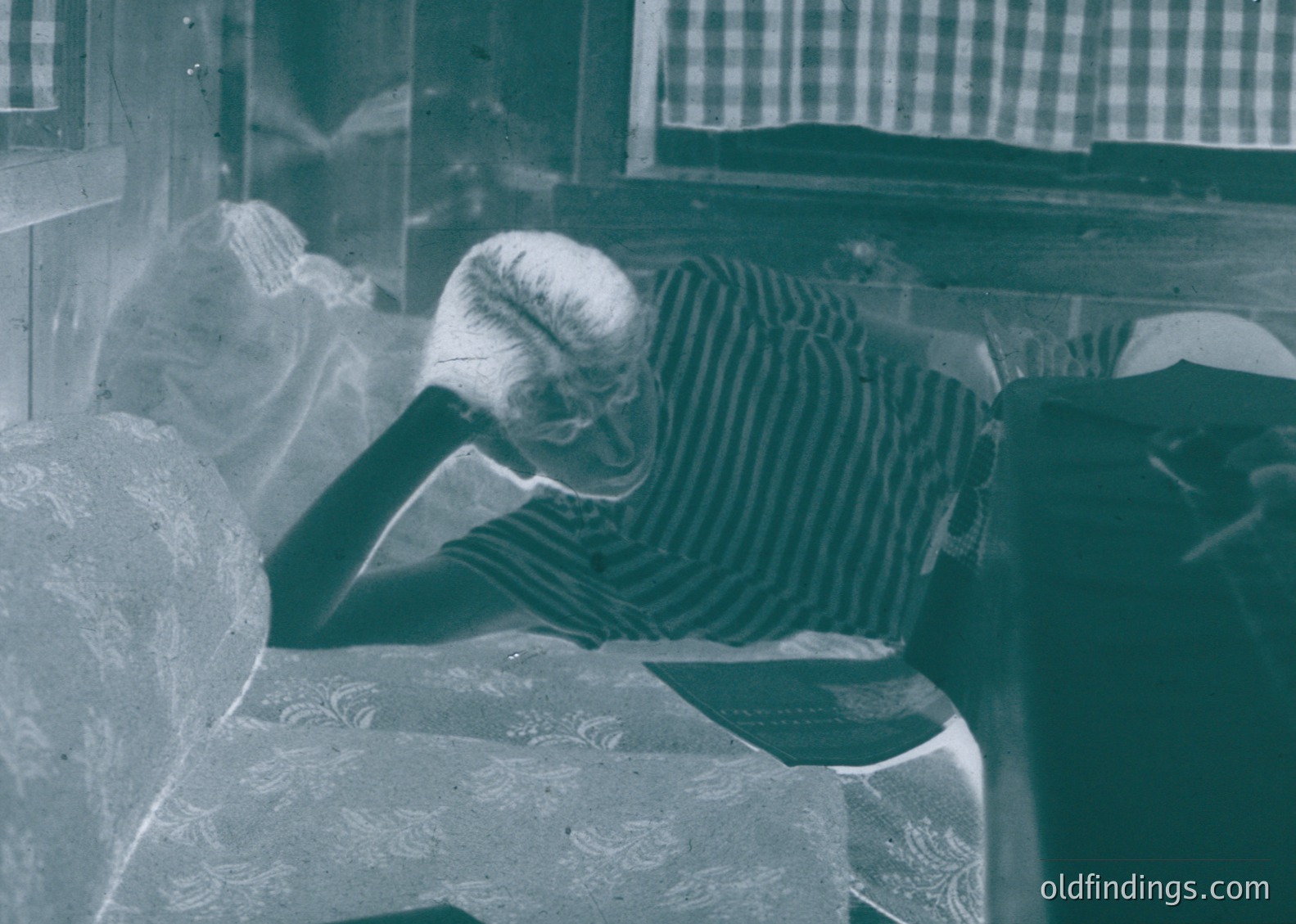 Vintage black-and-white portrait of an elderly woman resting her head on a marble or stone surface, likely a window ledge or sill. She wears a striped shirt and sits in a room with a checkered curtain and patterned wallpaper. Mid-20th century domestic interior, possibly .