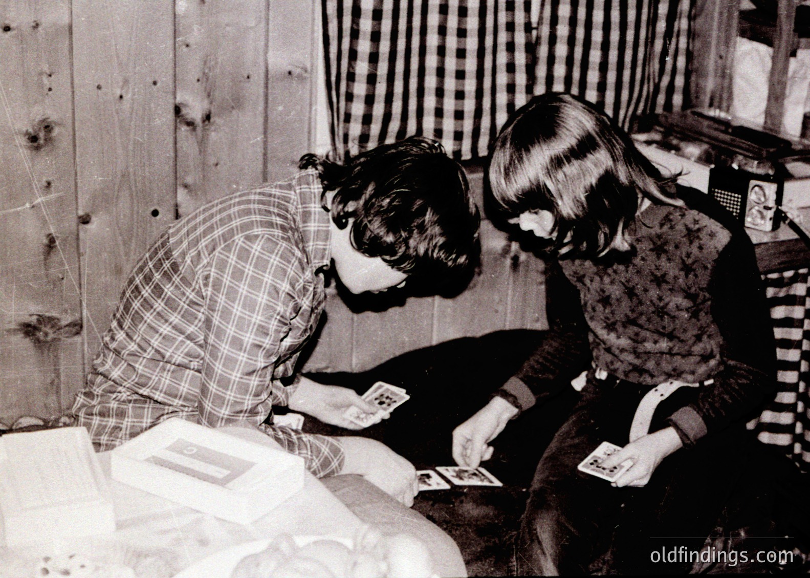 Two children playing cards on a couch in a modest, wood-paneled interior, likely mid-20th century. The woman’s checkered blouse and children’s clothing suggest 1950s–1960s American suburban life. Card game cards and a small envelope on the table indicate a casual, everyday moment.