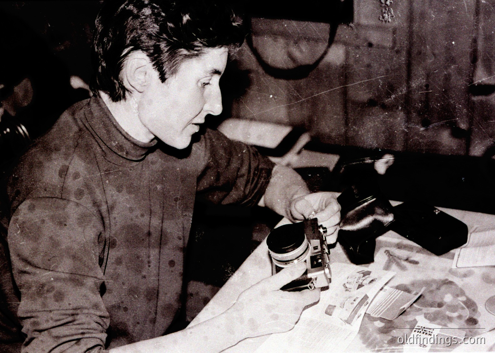 Black-and-white photo of a person meticulously assembling a small mechanical device, likely a camera or film projector, using tools and parts on a table. Mid-20th century workshop setting with scattered components and papers.