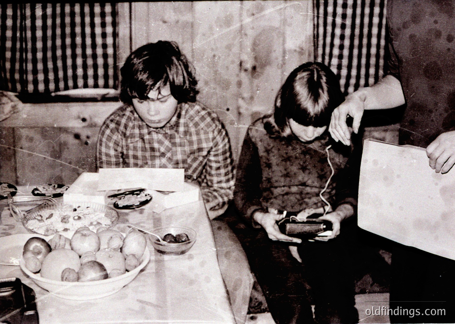 Two children in 1970s-era clothing—plaid shirt and glasses, sweater—engage with a vintage tape recorder and open notebook at a cluttered wooden table. Fresh fruit (apples, pears) and a bowl of shellfish sit beside them in a modest indoor setting. Likely Eastern Bloc region, mid-century domestic scene.