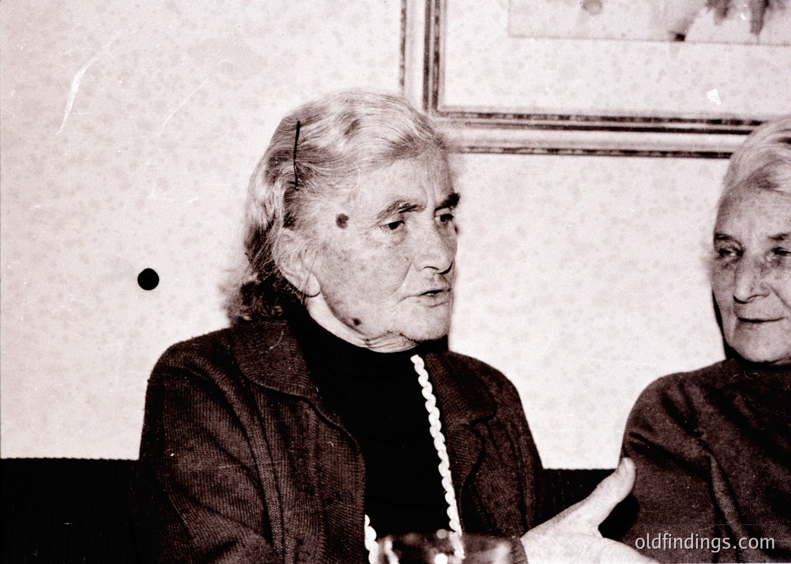 Black-and-white portrait of two elderly women in a formal indoor setting, likely mid-20th century. The woman in focus wears a structured blazer and pearl necklace, seated with a contemplative expression. Background includes framed artwork and a plain wall, suggesting a cultural or institutional venue.