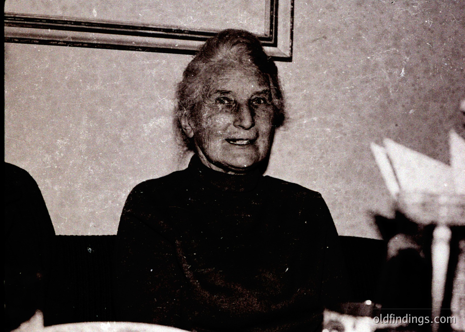 Portrait of an elderly woman in formal attire, likely mid-20th century. She wears a dark, high-necked dress with a pearl necklace and her hair styled in an updo. Background suggests an indoor setting with blurred figures, possibly a social gathering or event. [Vintage portrait ]