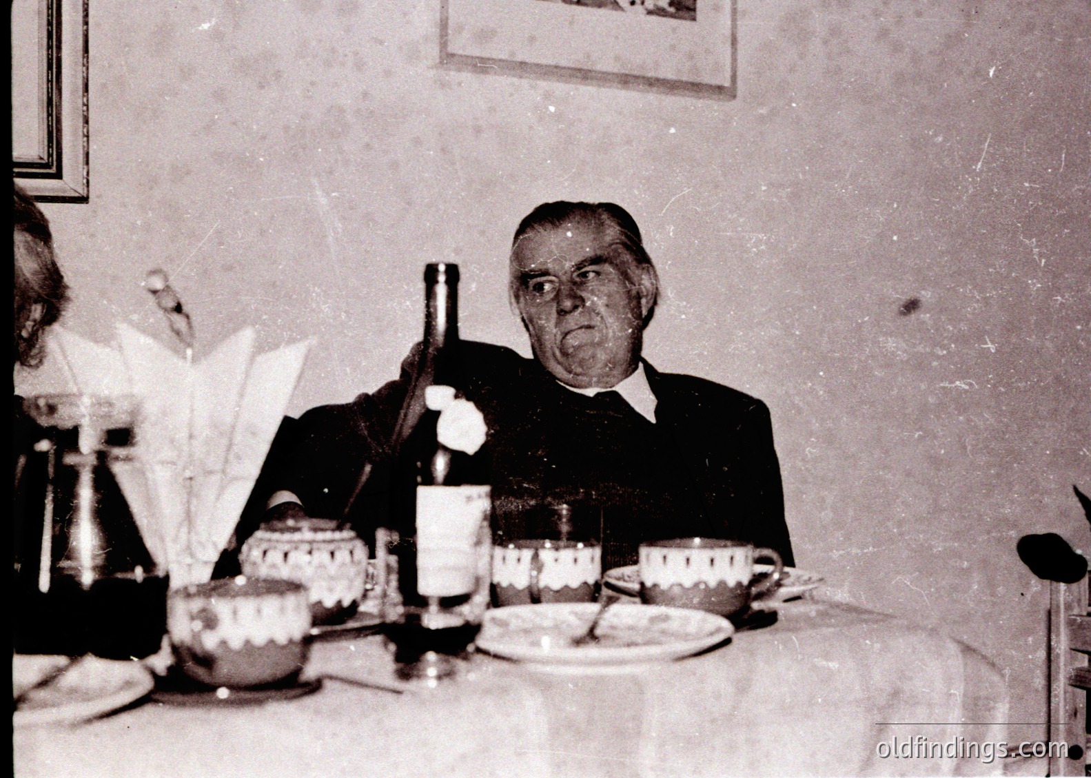 Mid-20th century indoor dining scene featuring an elderly man in formal attire holding a wine bottle. Table set with vintage china, a napkin holder, and a teapot. Plain wall and minimal decor suggest a modest, possibly rural setting.