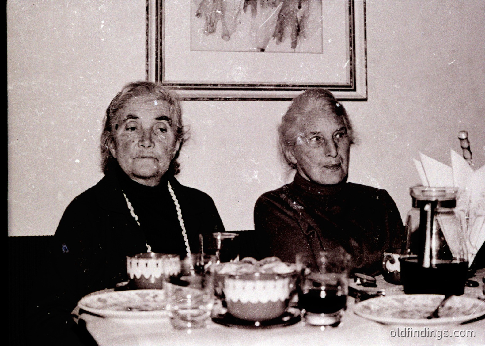 Two elderly women seated at a formal table, likely mid-20th century (1950s–1960s). Table setting includes vintage china, a teapot, and a framed artwork depicting a castle or fortress. Women wear high-neck dresses with pearl necklaces, suggesting a formal or commemorative occasion.