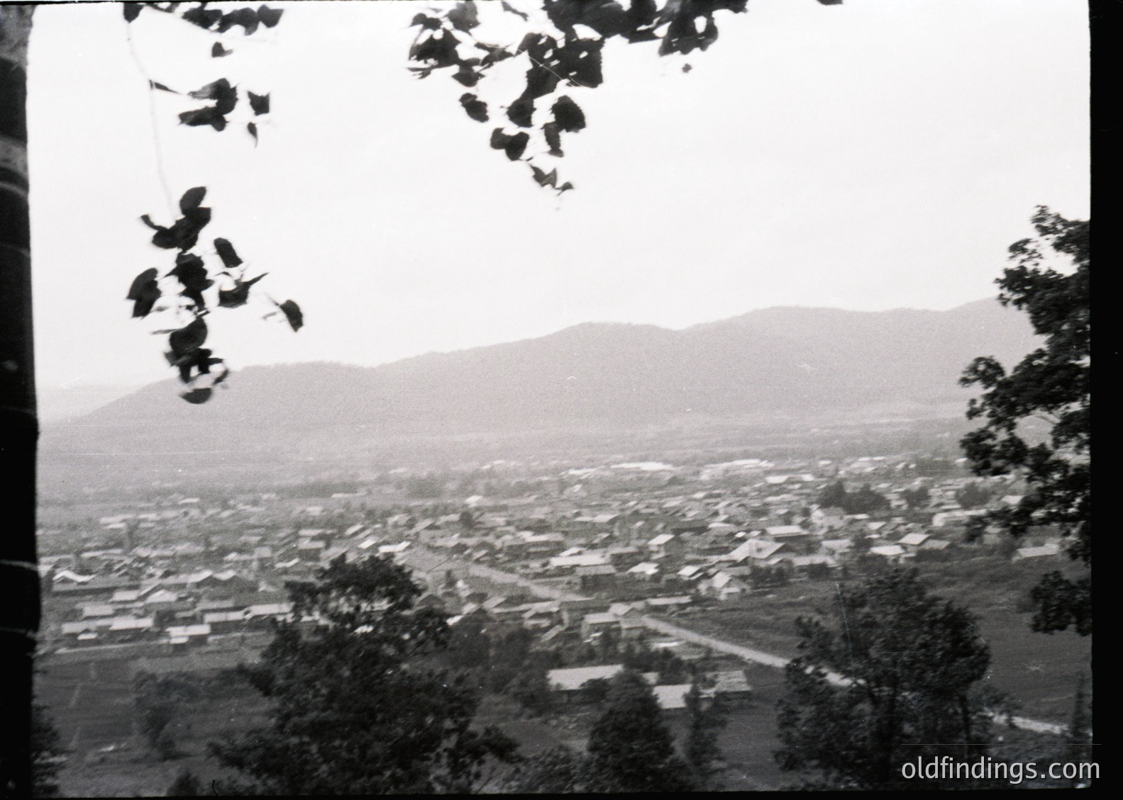 Aerial black-and-white view of a densely packed village nestled between rolling hills, framed by tree branches. Low-rise buildings with flat roofs and clustered layouts suggest a rural or early-industrial setting. Mist or haze obscures distant mountains, adding depth. Likely mid-20th century based on architectural style and monochrome quality.