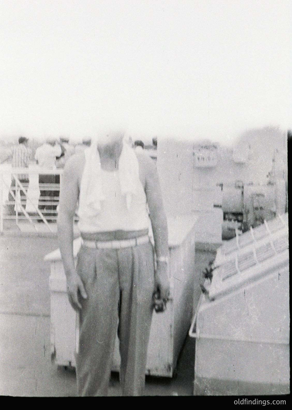 Mid-20th century man in loose trousers and tank top stands on a dock, holding a small object. Industrial port setting with visible cranes and ships in background. Likely 1950s–1960s, possibly Eastern Europe or USSR-era port.