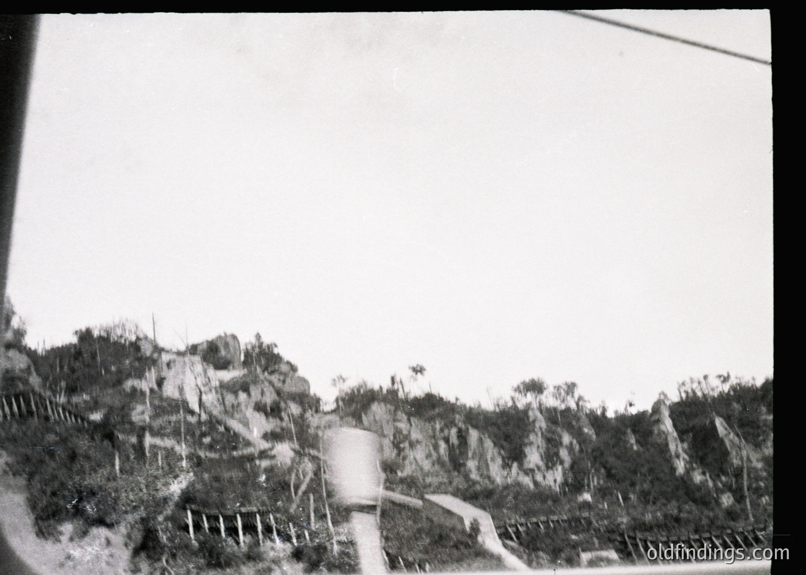 Black-and-white rural landscape featuring a dirt road winding through rocky terrain with sparse vegetation. A cylindrical water tank and wooden fence frame the scene, suggesting agricultural or remote settlement. Likely mid-20th century due to photographic style.