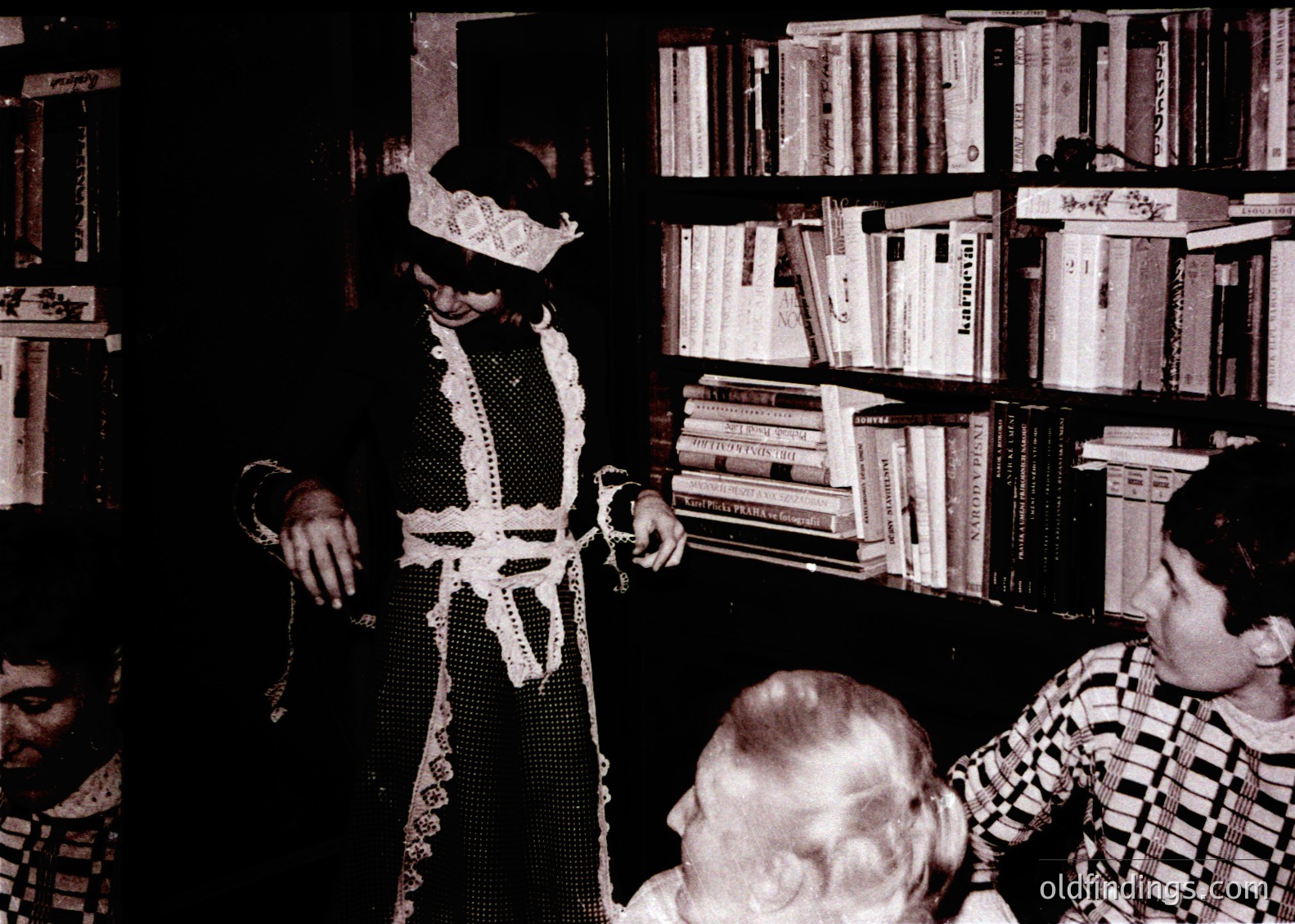 Vintage black-and-white photo of a theatrical performance in a dimly lit room. Central figure wears a dramatic, lace-trimmed costume with a wide-brimmed hat, holding a prop. Audience sits in casual attire, engrossed. Bookshelves filled with hardcover volumes line the background, suggesting a literary or cultural event. Likely mid-20th century, possibly 1950s–1960s.