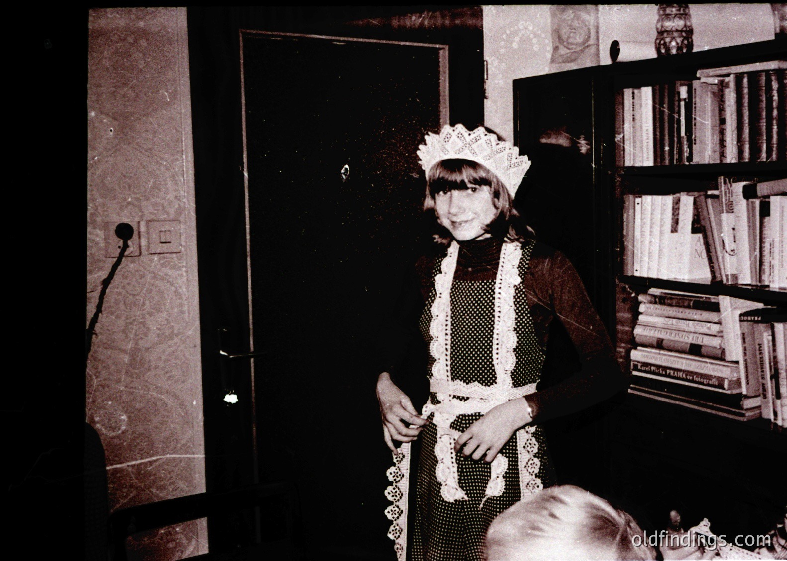 Vintage black-and-white photo of a young girl in a maid’s uniform—lace apron, blouse, and cap—posing indoors beside a bookshelf. Mid-20th century domestic setting, likely 1950s–1960s. Wallpaper and vintage decor suggest a mid-century home.