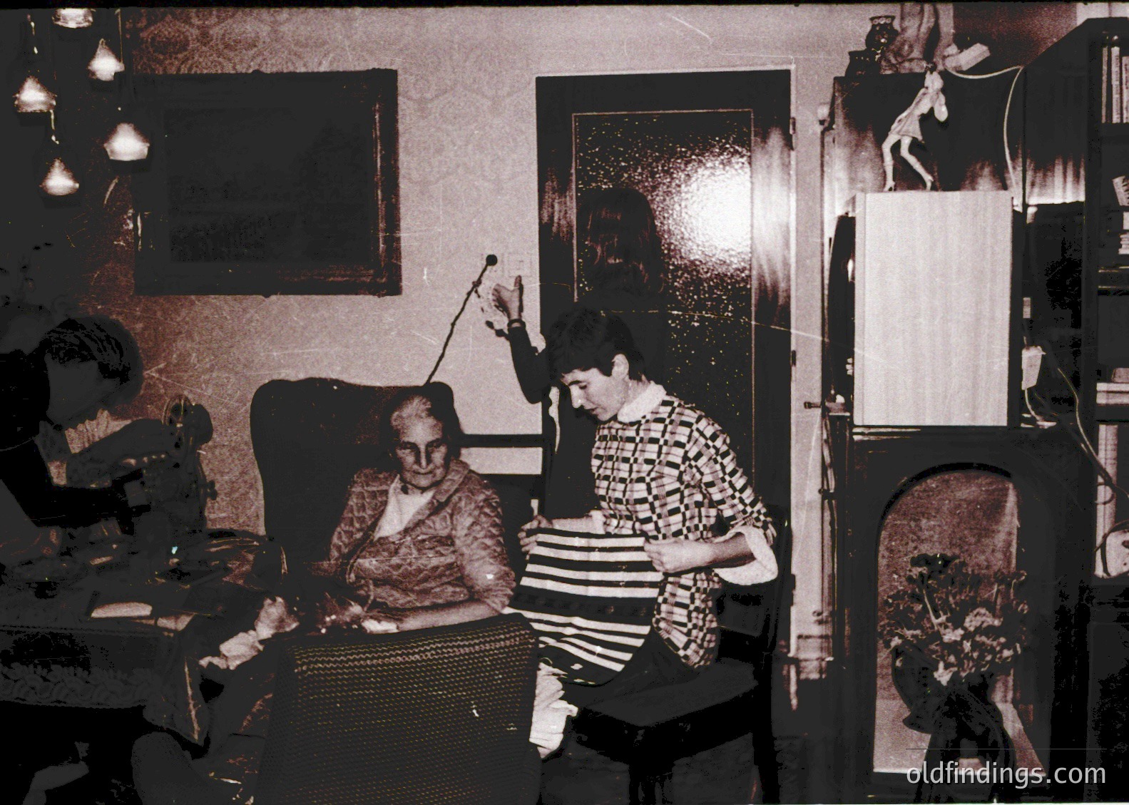 Vintage sepia-toned indoor portrait of two women in a modest living space, likely 1960s–1970s. One woman, seated on a woven chair, holds a cane; the other, in a striped sweater, gestures with her hand. Decor includes framed art, a curtained window, and a door with a carved relief. Warm lighting from a chandelier enhances the intimate setting.