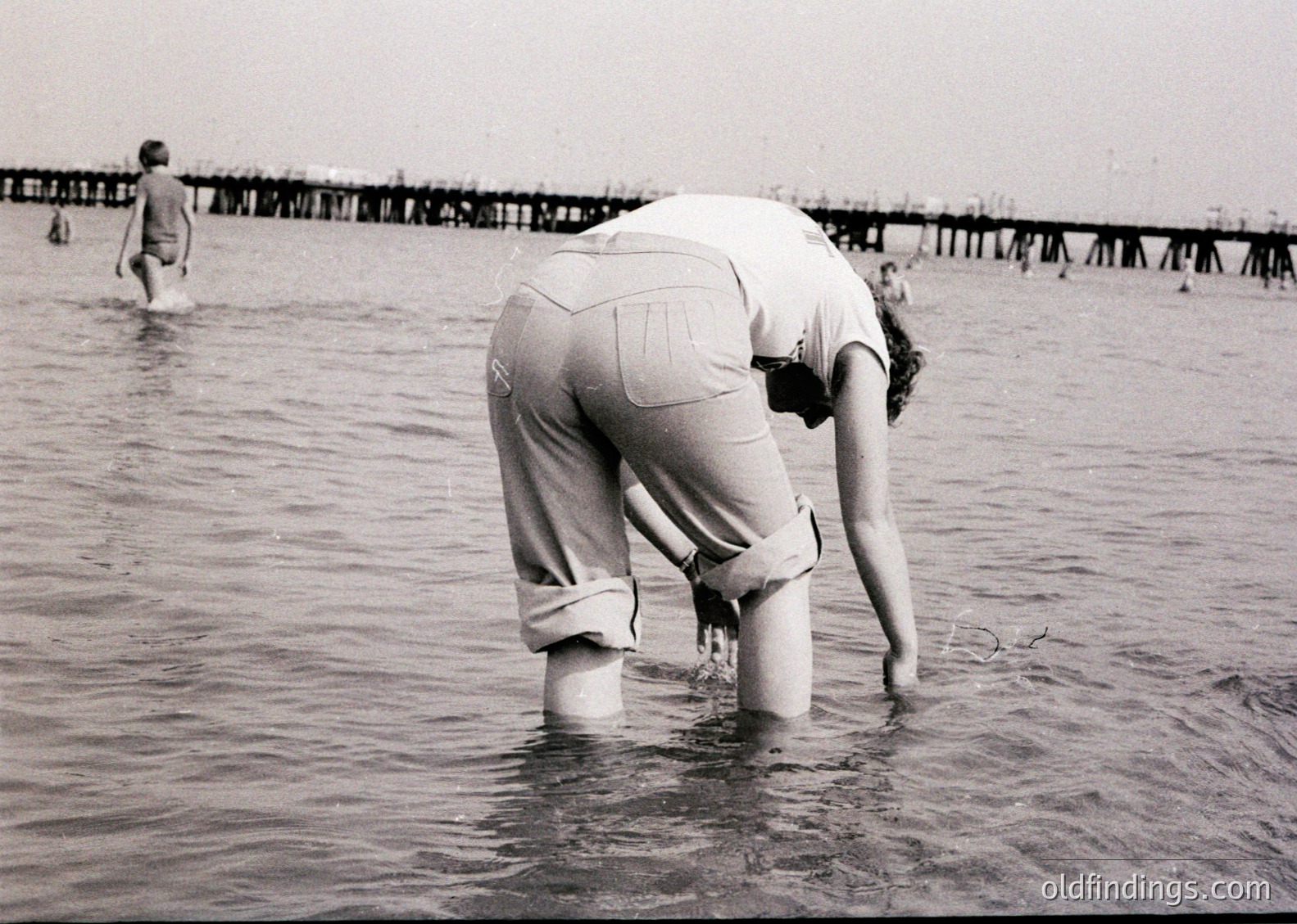 Black-and-white beach scene from mid-20th century (1950s–1960s), showing women in knee-length swimsuits wading in shallow water. Wooden pier and wooden walkway with railings in background. Casual, retro coastal lifestyle captured.