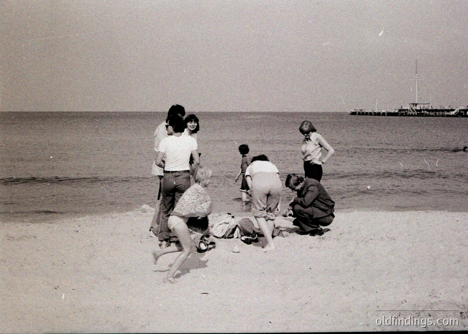 Group of people on a sandy beach, mid-20th century. One person lies on sand while others kneel or stand around, likely during a seaside outing or picnic. Distant sailboat visible in background.