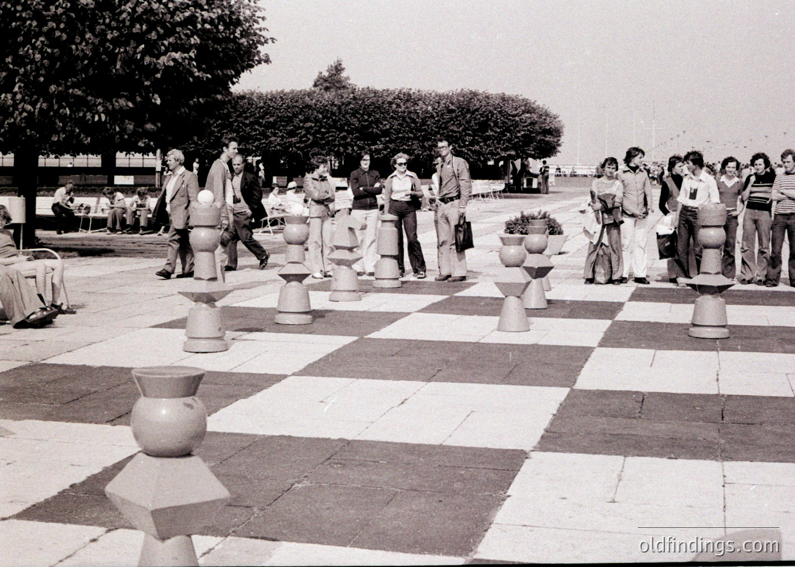 Large-scale outdoor chess game on a checkerboard-patterned plaza with ceramic chess pieces. Mid-20th century attire suggests or . Group of players engaged in casual competition, likely at a public event or festival. Urban setting with trees and greenery in background.