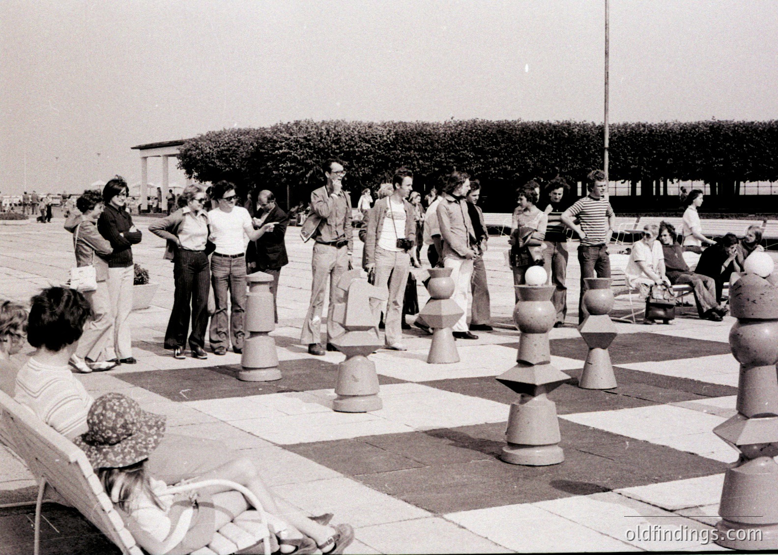 Large outdoor chess set with 1970s-style attire; people engaged in casual play. Urban park setting with trees and paved area.
