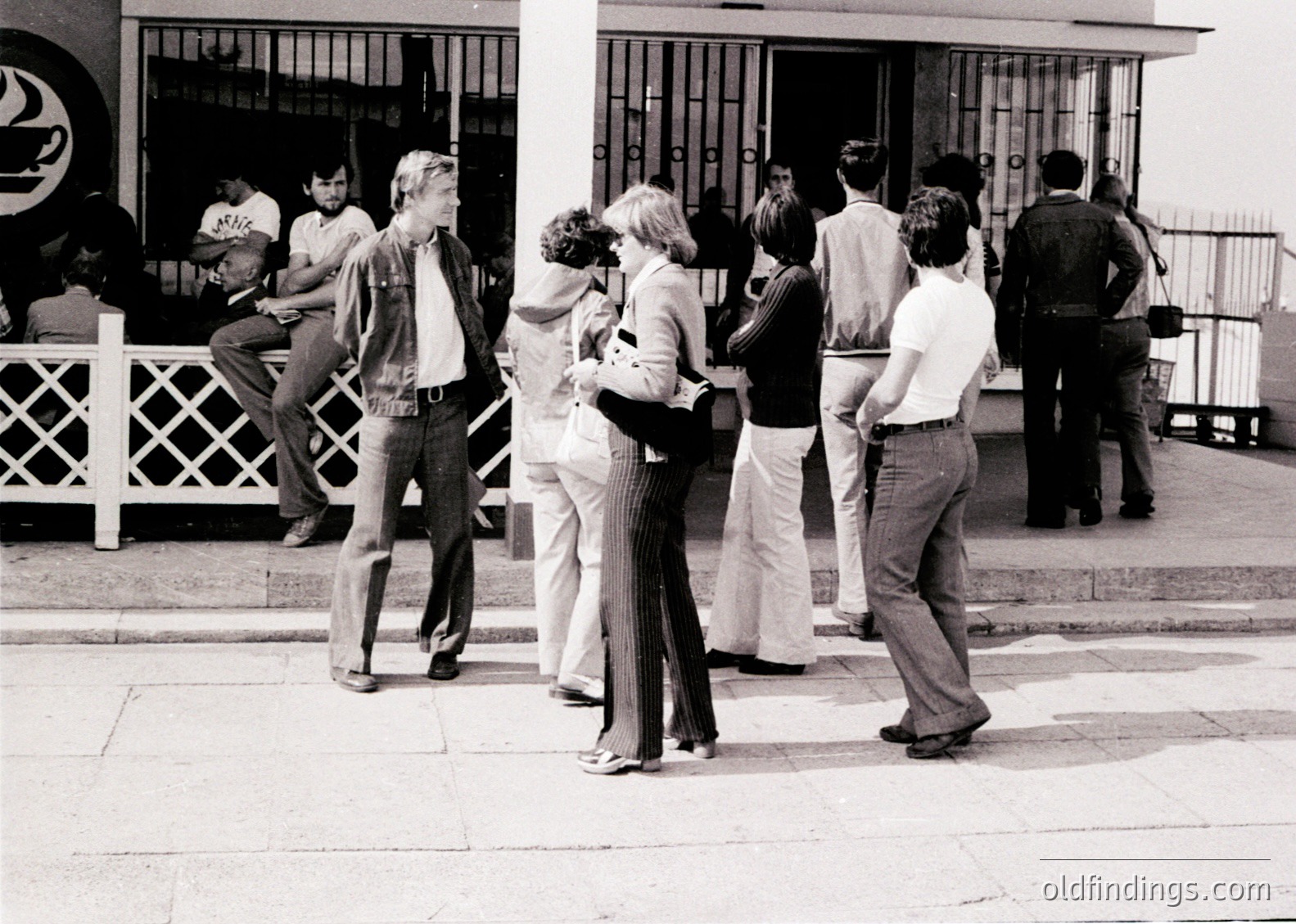 Group of young adults in 1970s streetwear—bell-bottoms, wide lapels, and casual poses—gathering outside a building with wrought-iron railings. Urban setting with mid-century concrete architecture. Likely Eastern European based on attire and signage.