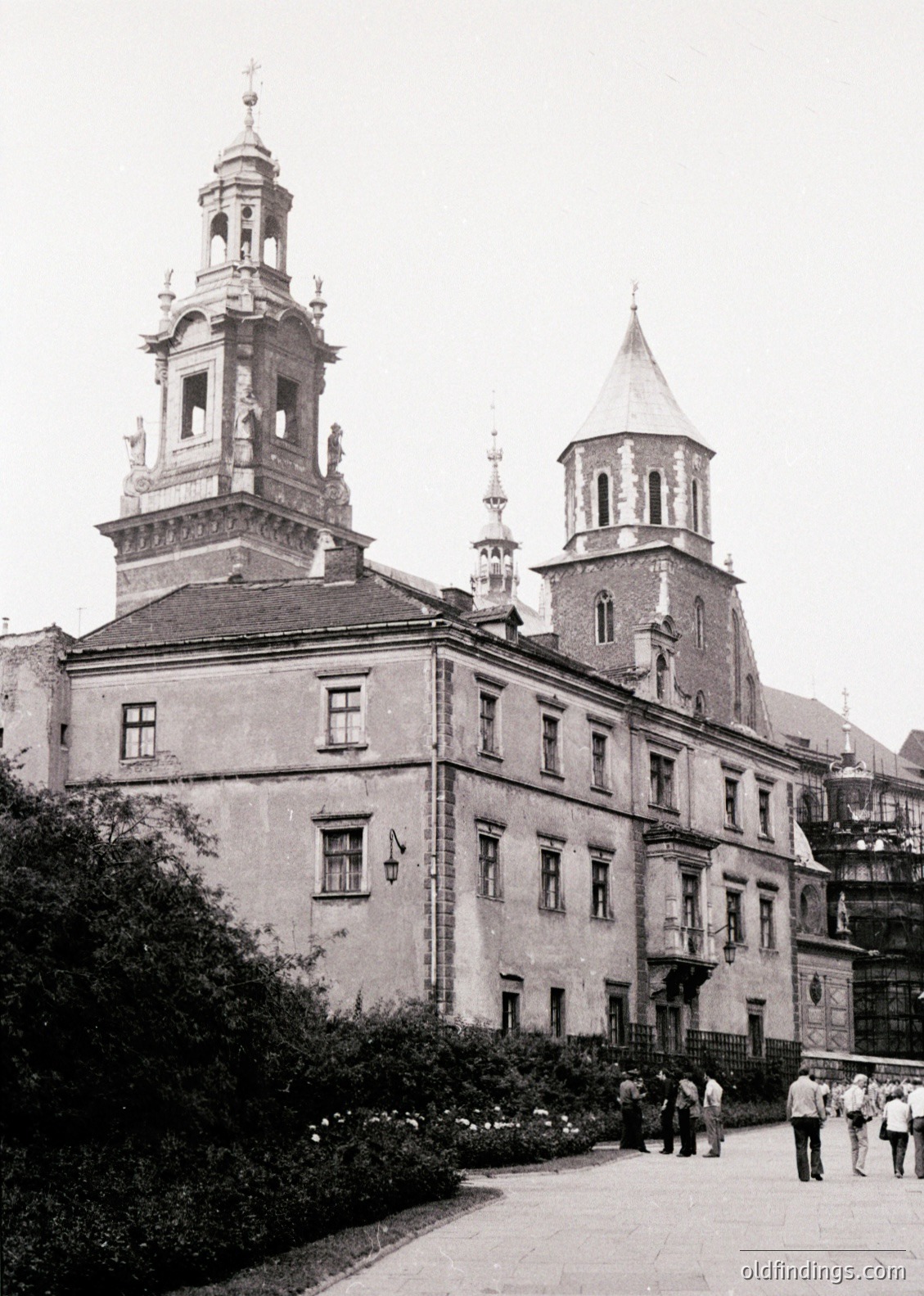 Historic Baroque-style building with twin towers and decorative spires, likely a 17th–18th century church or palace. Symmetrical facade features arched windows and ornate stonework. Courtyard entrance flanked by shrubs; small group of people in mid-20th-century attire. Potential location: Central/Eastern Europe.