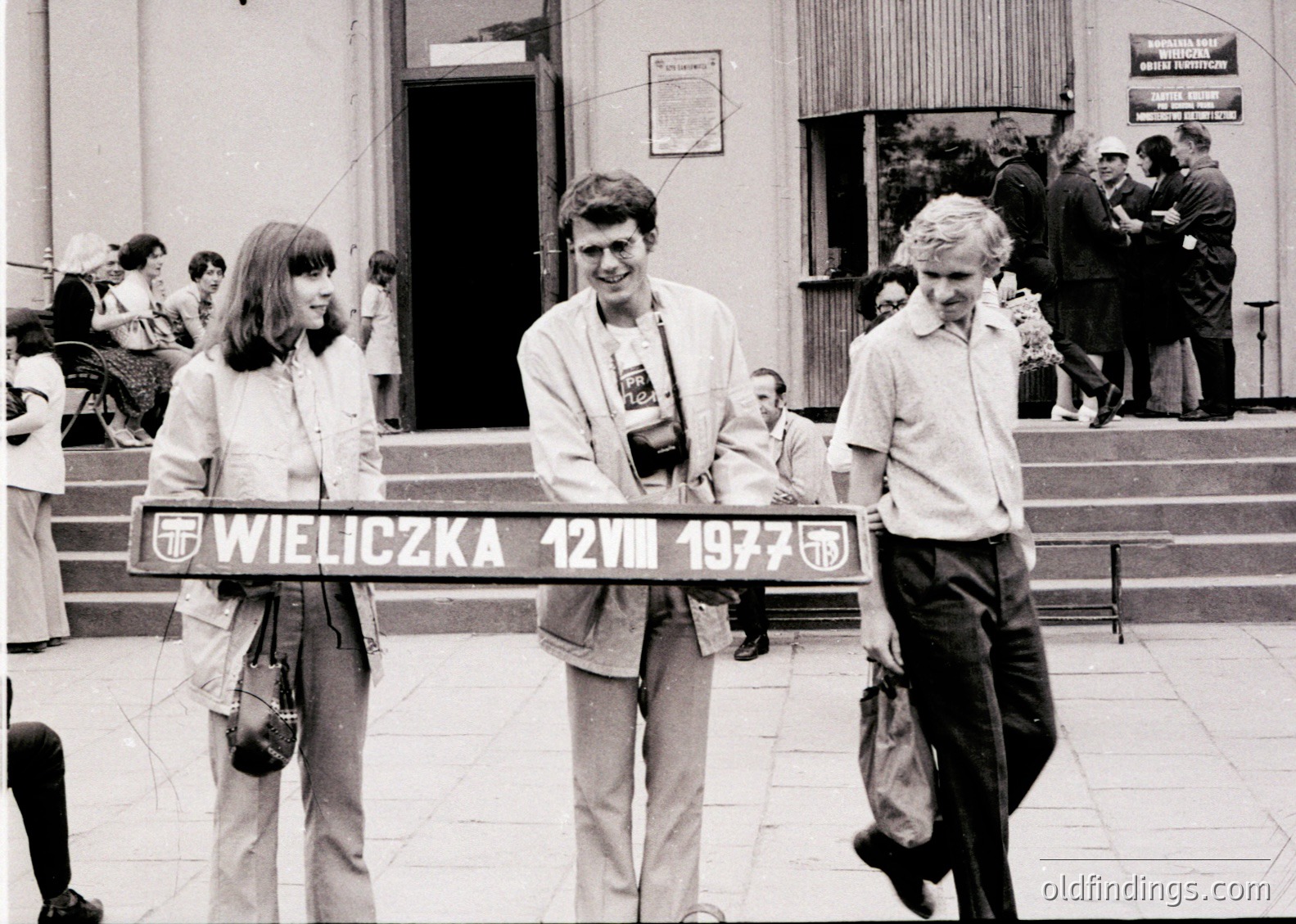 Black-and-white snapshot from **Wieliczka Salt Mine, Poland, 1977**. Three tourists pose with a sign marking the date and location, surrounded by crowds in formal attire. Classic 1970s fashion and Soviet-era architecture visible. Ideal for historical research, travel archives, or vintage tourism references.