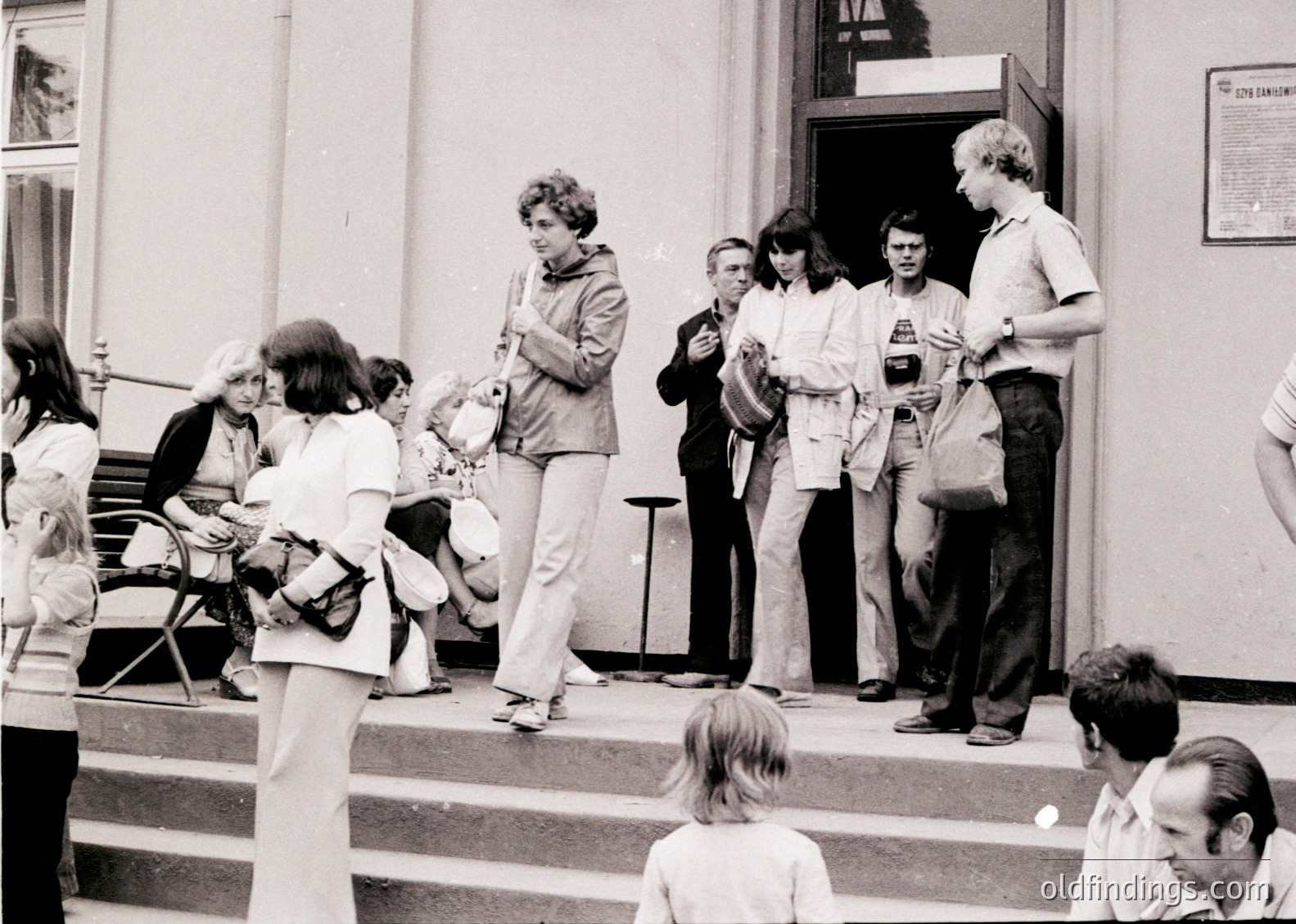 1970s street scene outside a Soviet-era building, featuring a diverse group of people in casual attire—men in button-downs, women in blouses and skirts, and a child in shorts. A woman in a light jacket holds a camera, while others stand or sit on steps. A sign with Cyrillic text suggests Eastern European location.