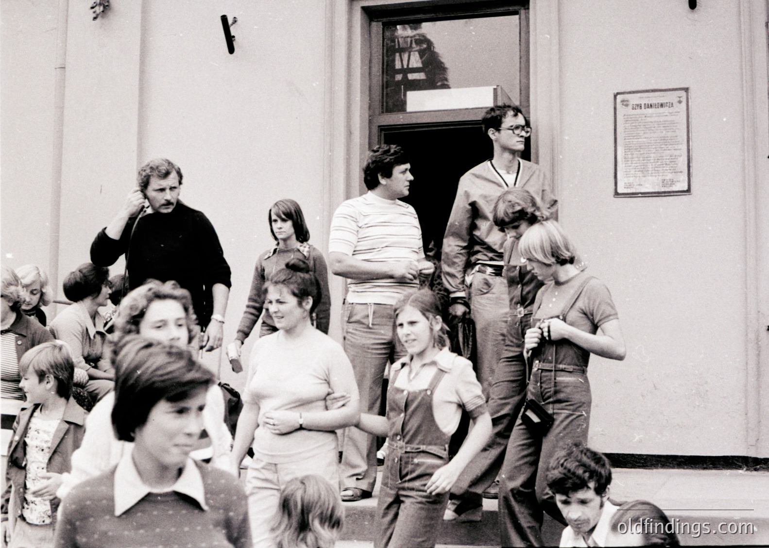 A candid black-and-white street scene from the **1970s**, featuring a diverse group of young adults and children gathered outside a building. Casual 70s fashion—bell-bottoms, striped shirts, and short hair—dominates. A sign on the wall reads *"Център на младежта"* (Youth Center in Bulgarian), suggesting a social hub in **Sofia, Bulgaria**. The composition captures spontaneous urban life and communal activity.