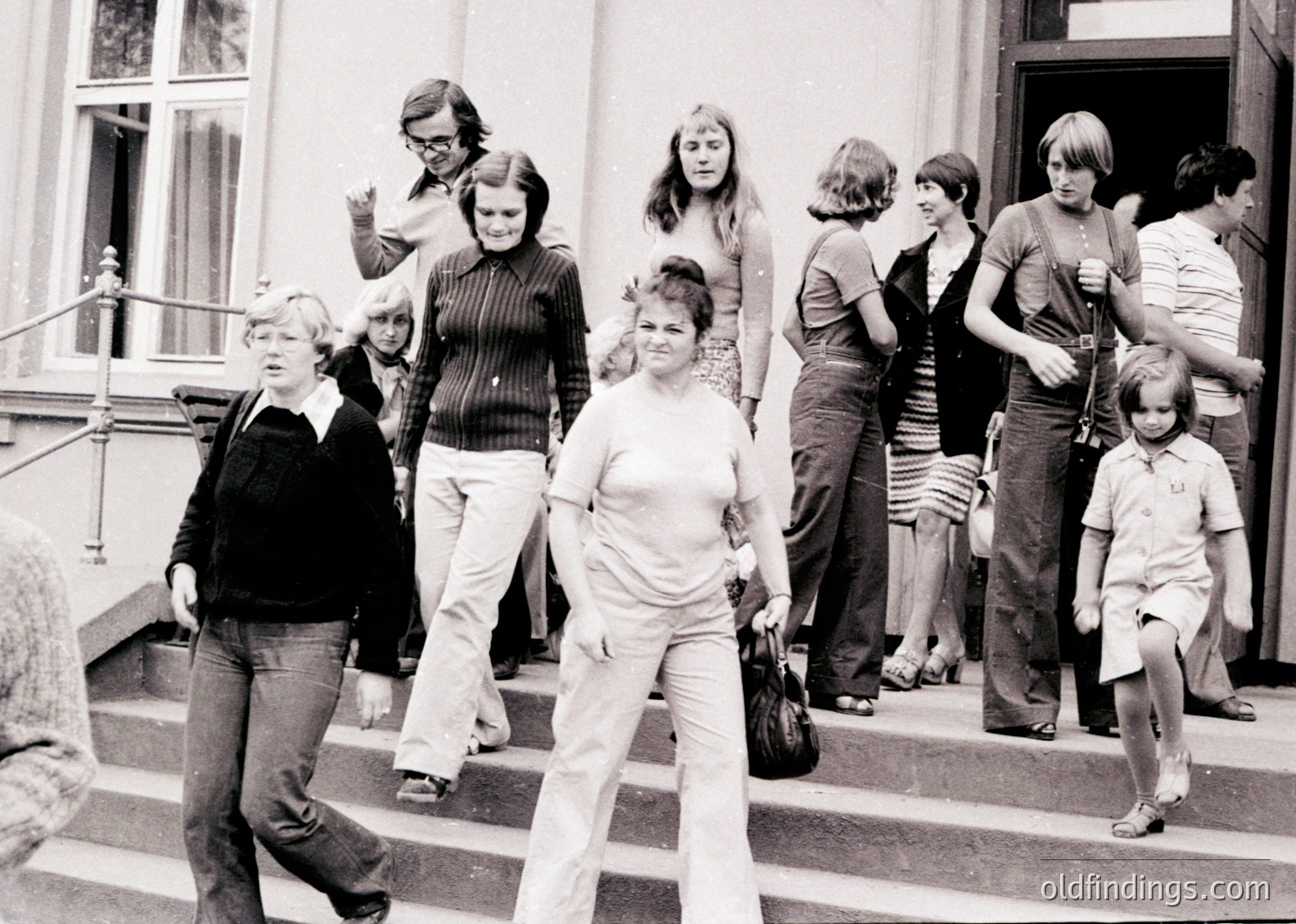 Group of 12 people descending grand stone staircase in 1970s European urban setting. Women in wide-leg trousers, men in short-sleeved button-ups; one child holding handrail. Architectural details: wrought-iron balcony, arched doorway, and light-colored facade.