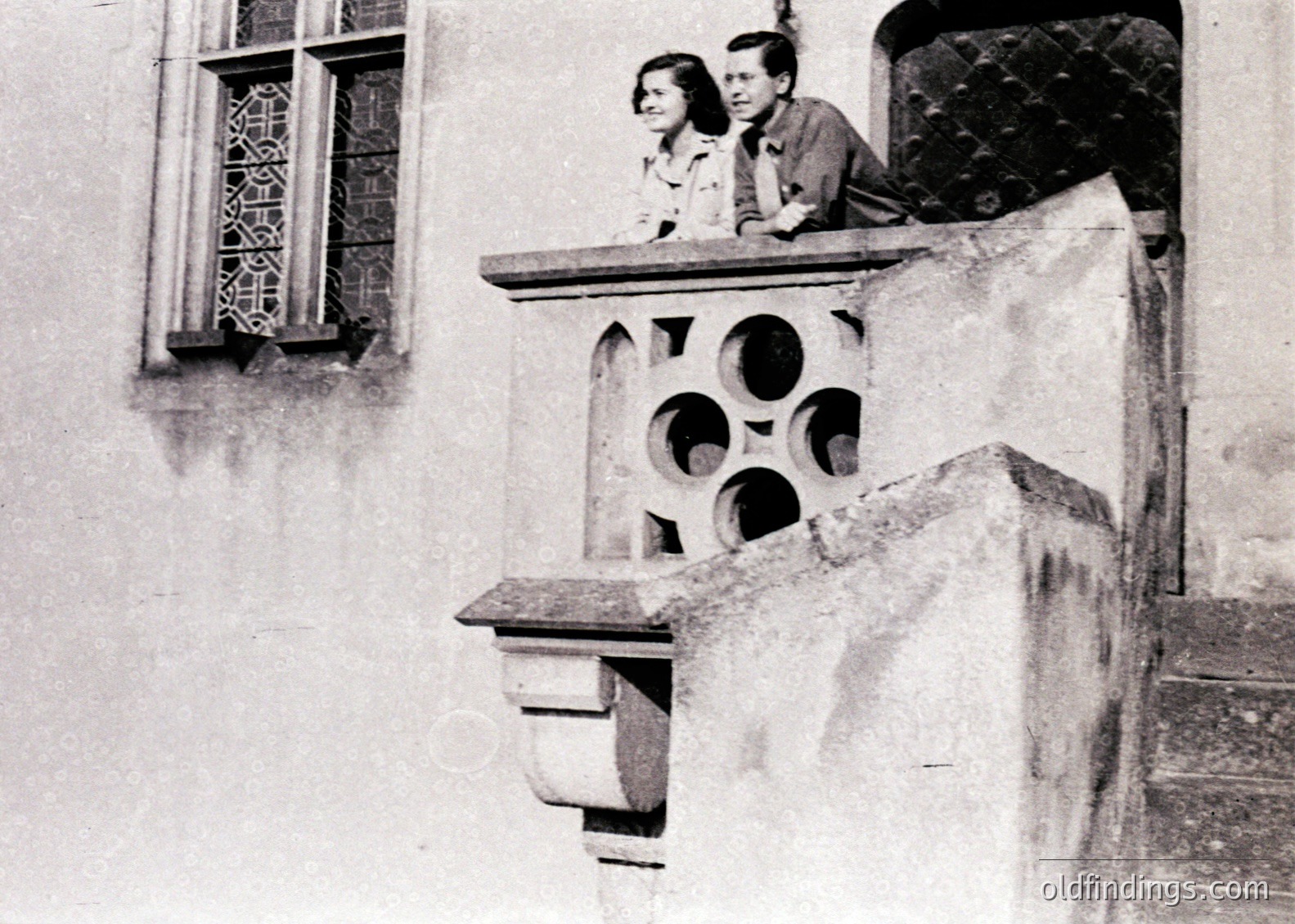 Black-and-white photo of two individuals leaning on a stone balcony with decorative circular openings, set against a building with ornate lattice windows. Mid-20th century European architectural style.