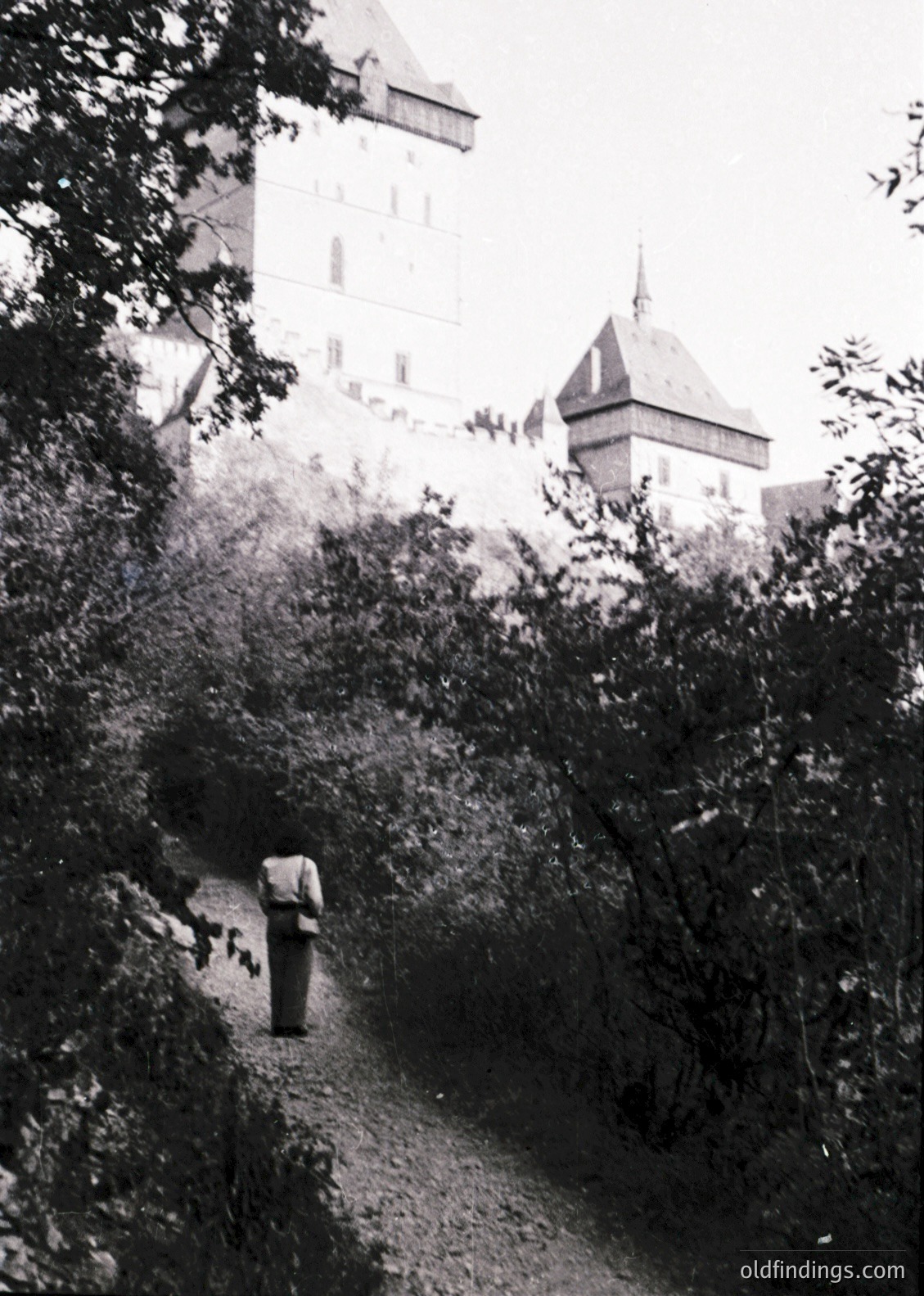 Medieval castle tower framed by dense foliage, likely a European fortress. A lone figure in period clothing walks a winding path, suggesting historical or vintage photography. Architectural details include steep gables and battlements.