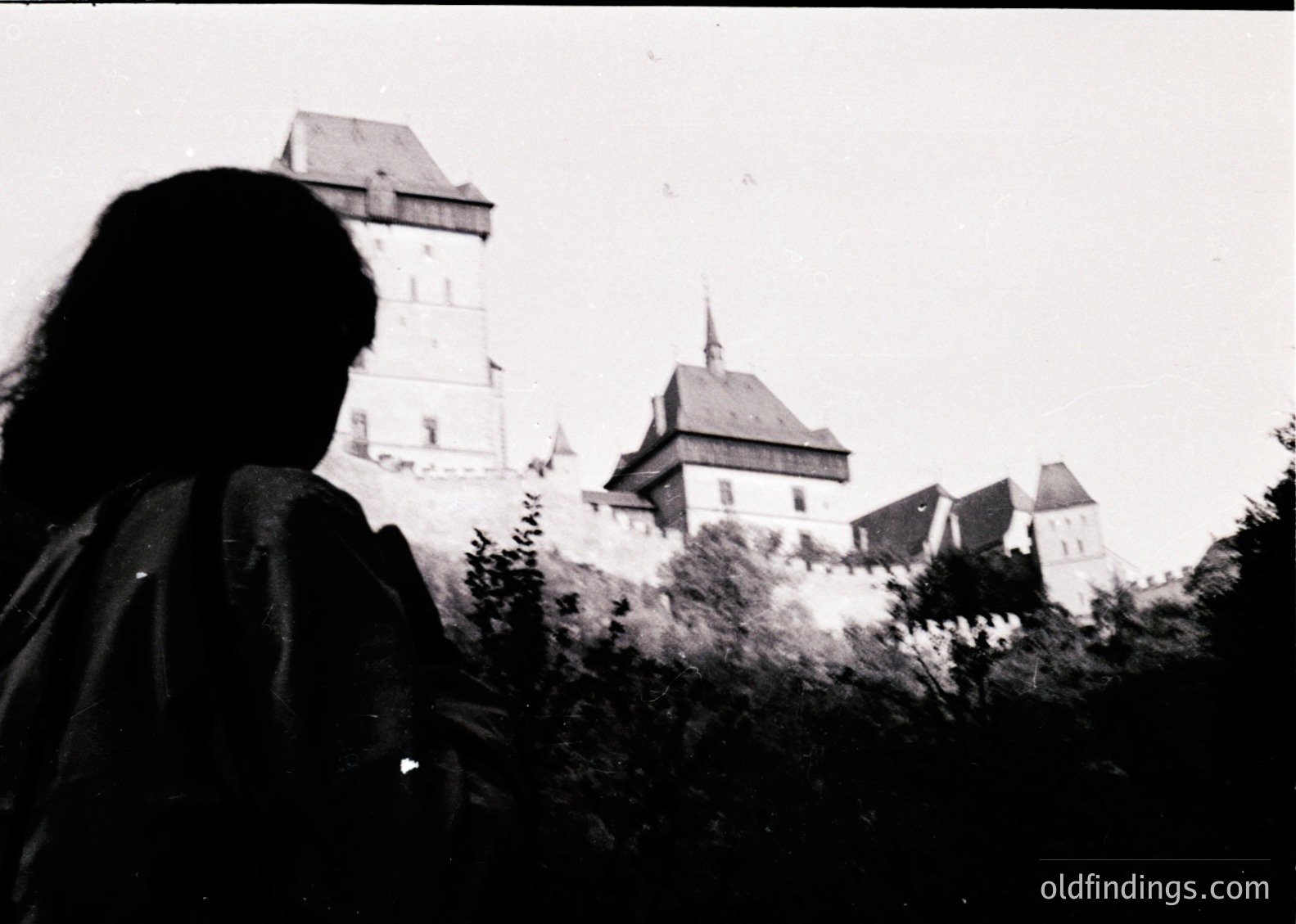 Silhouetted figure frames medieval castle with steep-turreted towers and gabled roofs, likely štejnCastle (). Black-and-white grain suggests – photography. Architectural details include crenellations and a central tower. Ideal for historical research or vintage design references.