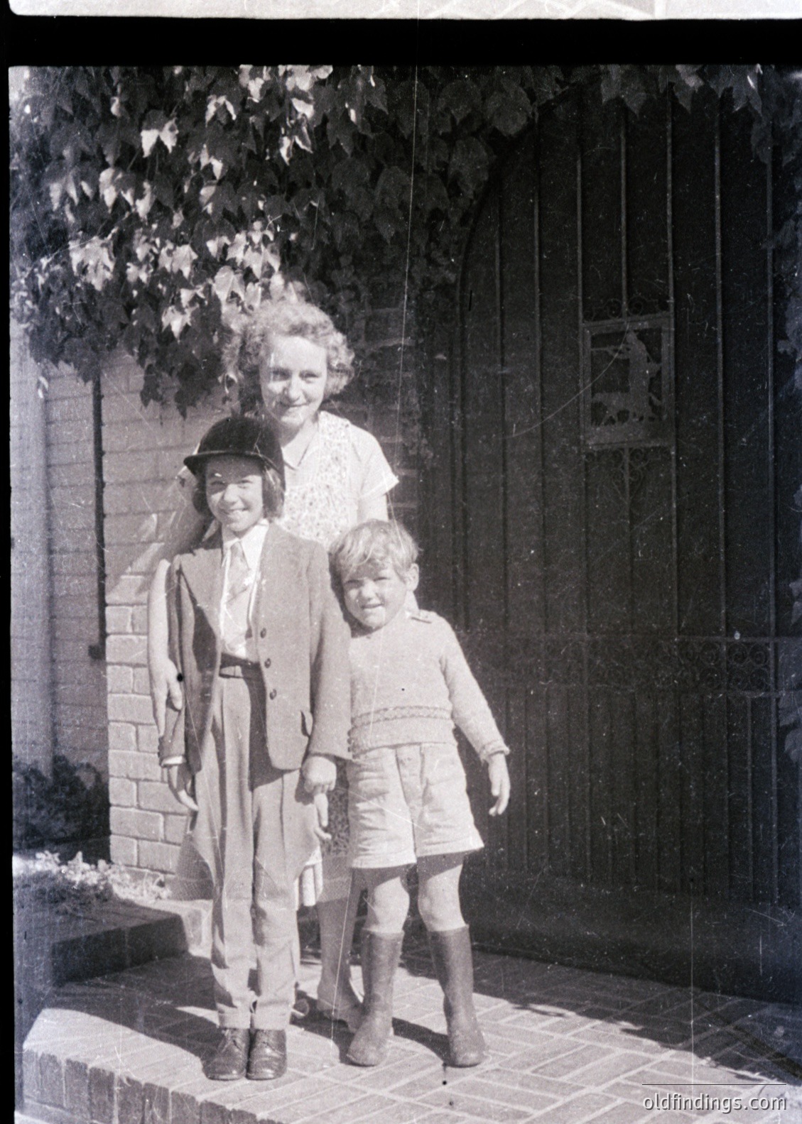 Vintage black-and-white family portrait featuring a woman in a high-necked blouse and a child in a sailor-style jacket with knee-high socks, standing beside a younger child in a sweater and shorts. Brick wall and wooden gate with a sign in the background suggest a residential setting, likely mid-20th century (1940s–1950s). Classic candid style ideal for historical research or nostalgic design references.