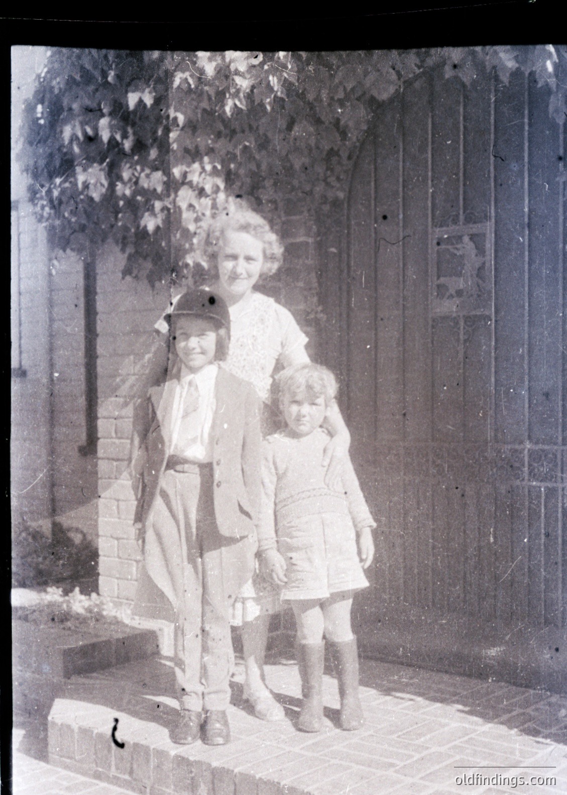 Vintage sepia-toned family portrait from early 20th century, likely 1910s–1920s. Woman in high-necked blouse and long skirt poses with two children—boy in sailor-style cap and girl in knee-length dress—standing on brick steps beside a wrought-iron gate. Decorative tile panel on gate features floral motifs. Residential setting suggests urban or suburban European/US context.