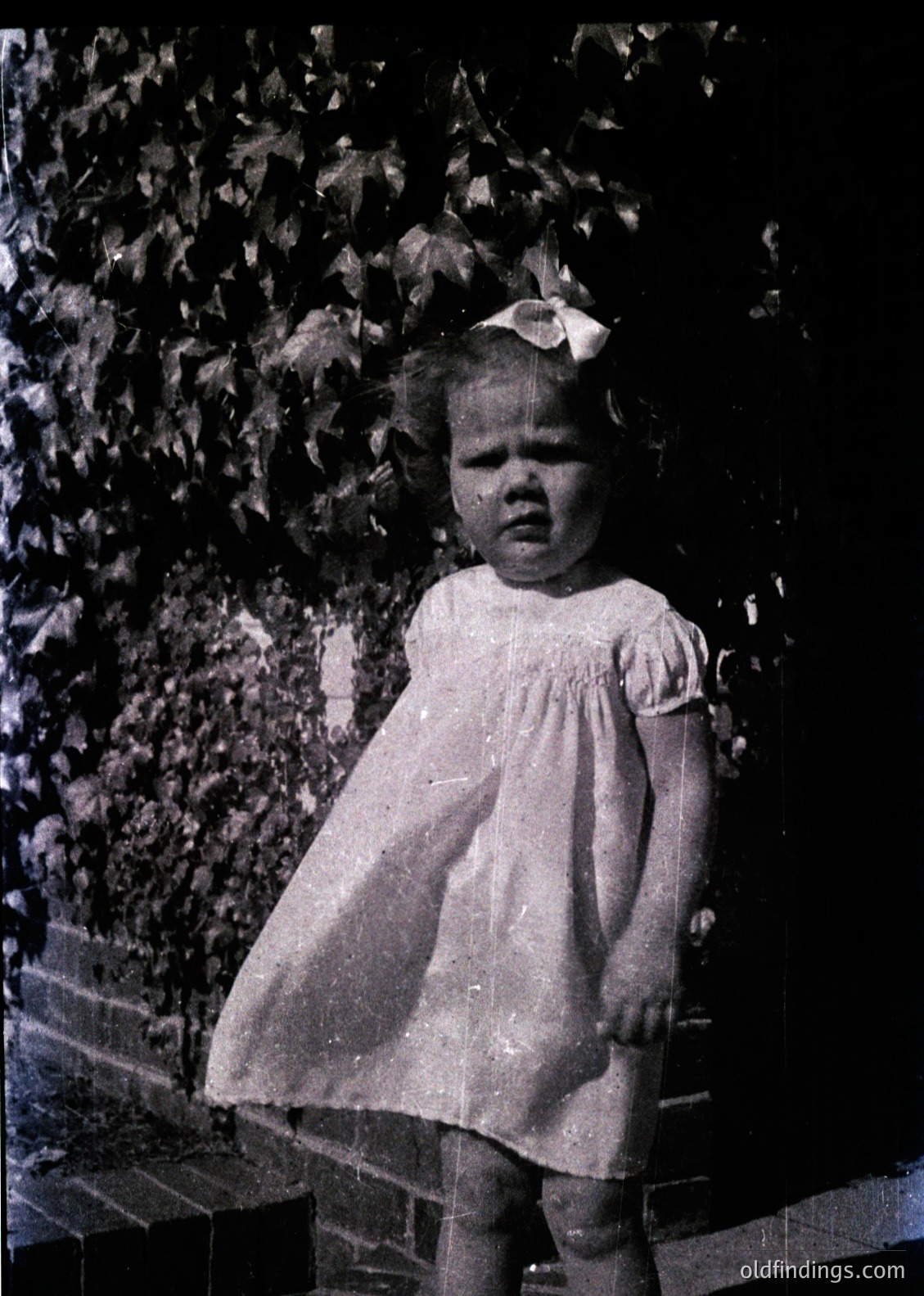 Vintage black-and-white portrait of a young girl in a 1950s-style dress with a bow, standing on stone steps beside leafy foliage. Her expression is neutral, framed by soft lighting.