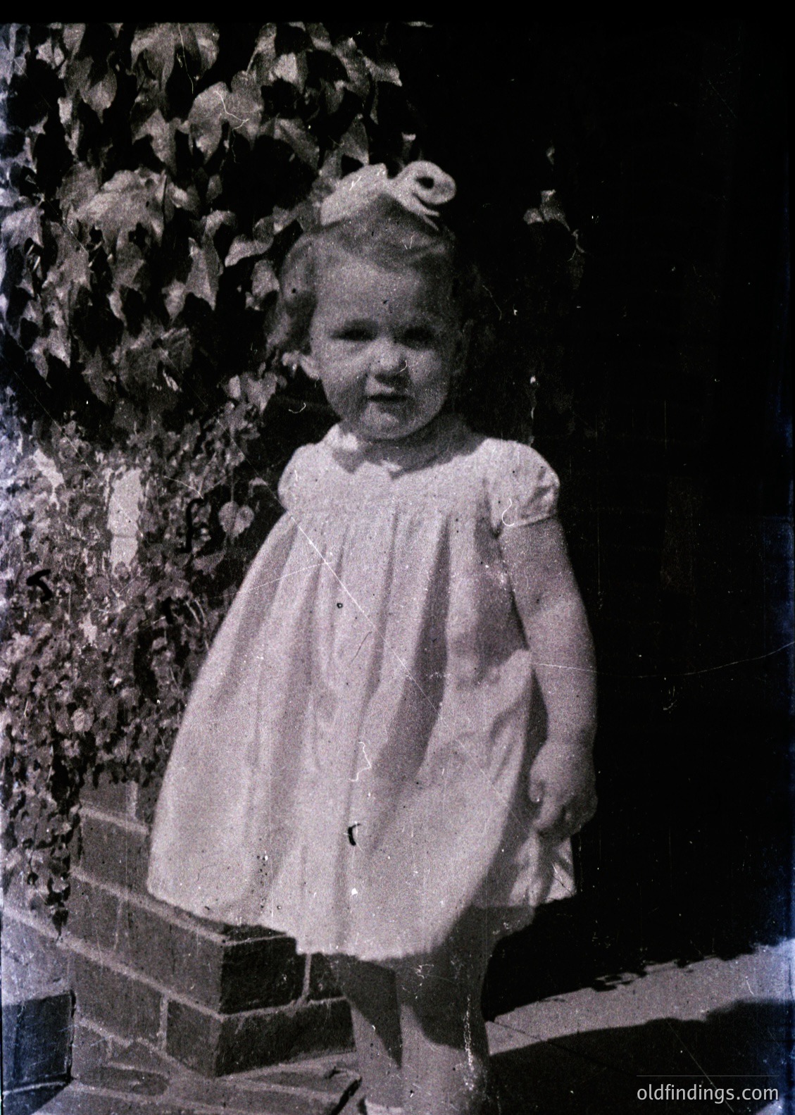 Vintage black-and-white portrait of a young girl in a 1950s-style dress with bow hair accessory, standing on brick steps beside lush greenery. Candid smile captures innocence and mid-century fashion.