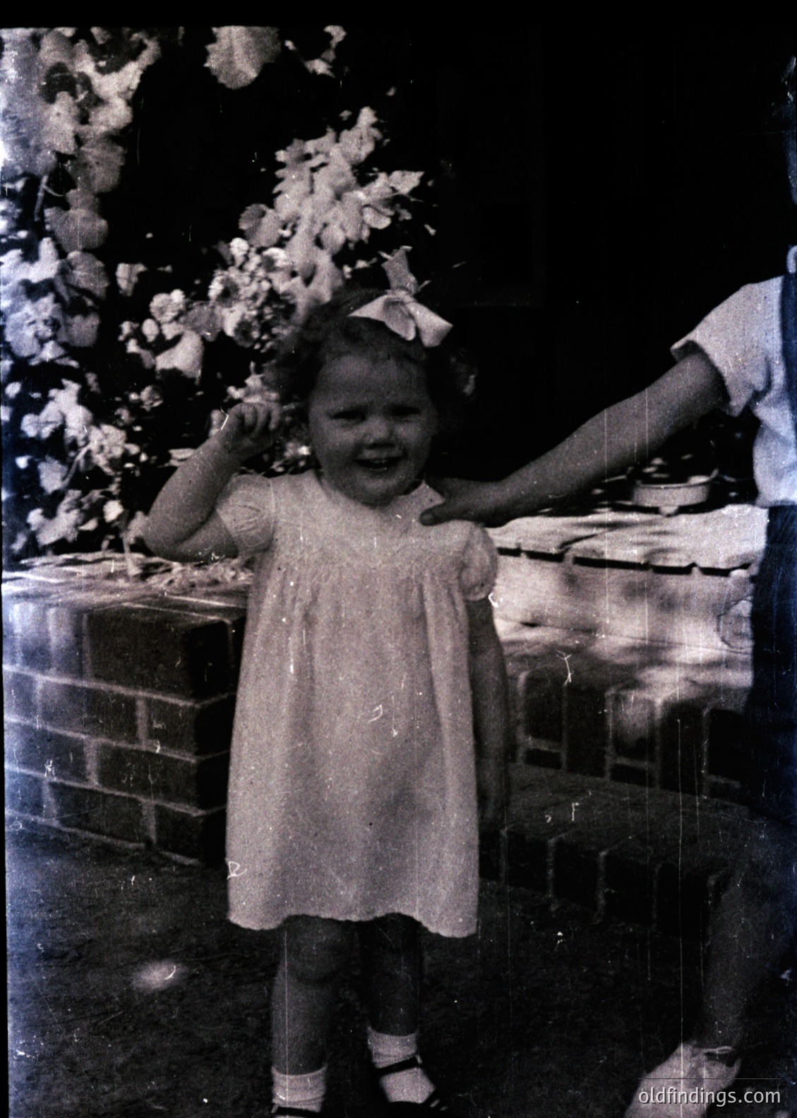 Vintage sepia-toned photo of a young girl in a short-sleeved dress with a bow, standing beside a brick oven or stove. Her joyful expression contrasts with the dark, textured background. Likely mid-20th century household scene.