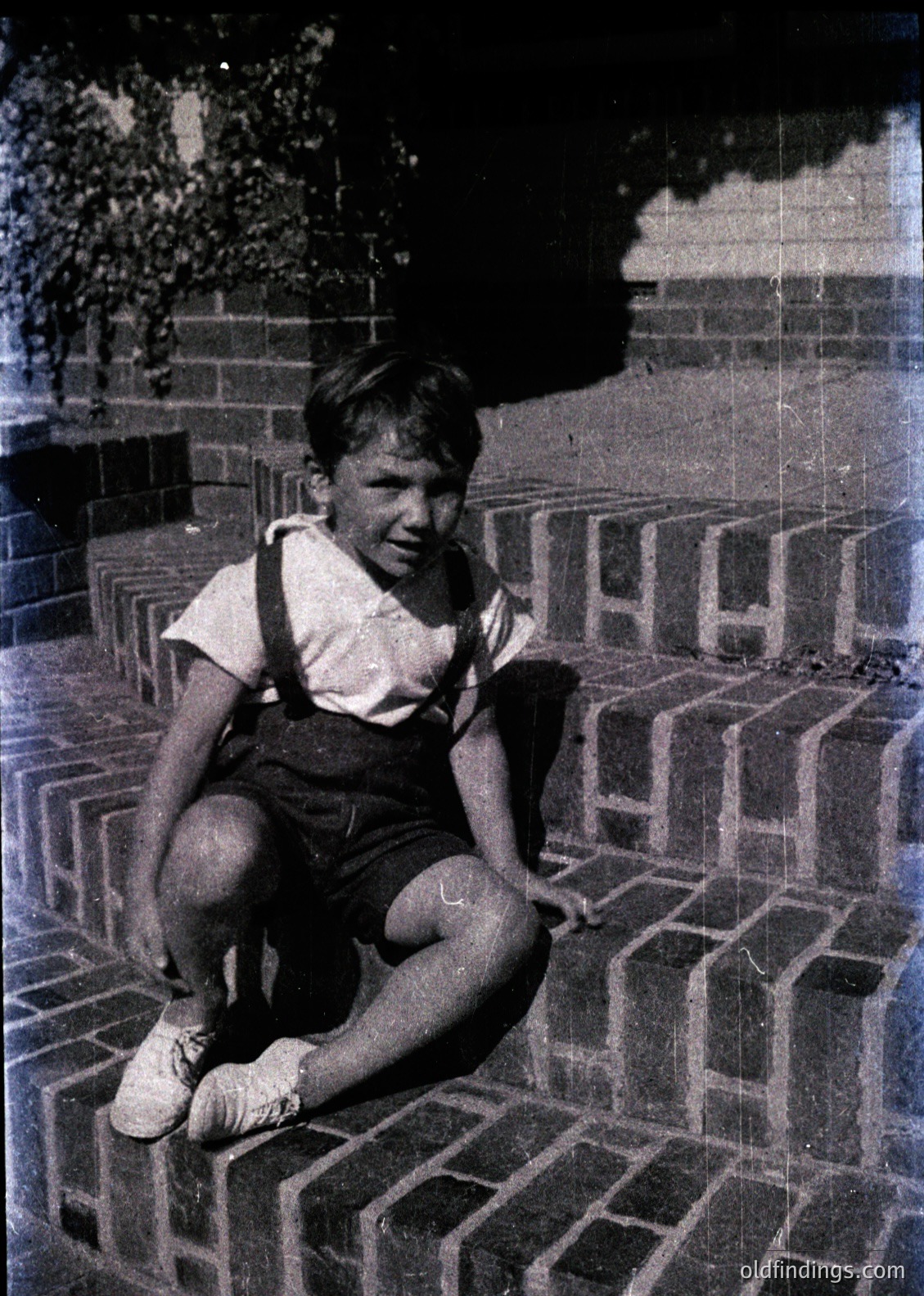 Vintage black-and-white photo of a boy in overalls and white shoes sitting on brick steps, smiling directly at the camera. Mid-20th century clothing and sepia tone suggest . Urban setting with brickwork and greenery in background.