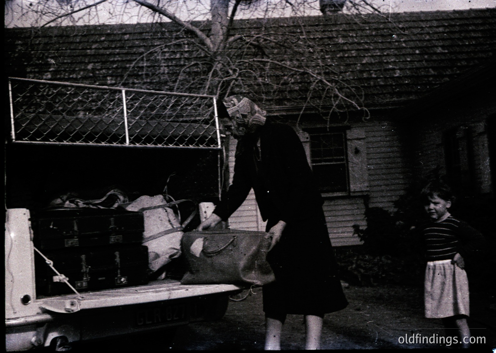 Mid-20th century black-and-white photo: Woman in dark coat and headscarf unloading a large basket from a vintage truck, while a child in striped dress observes. Brick courtyard with wooden fence and utility boxes.