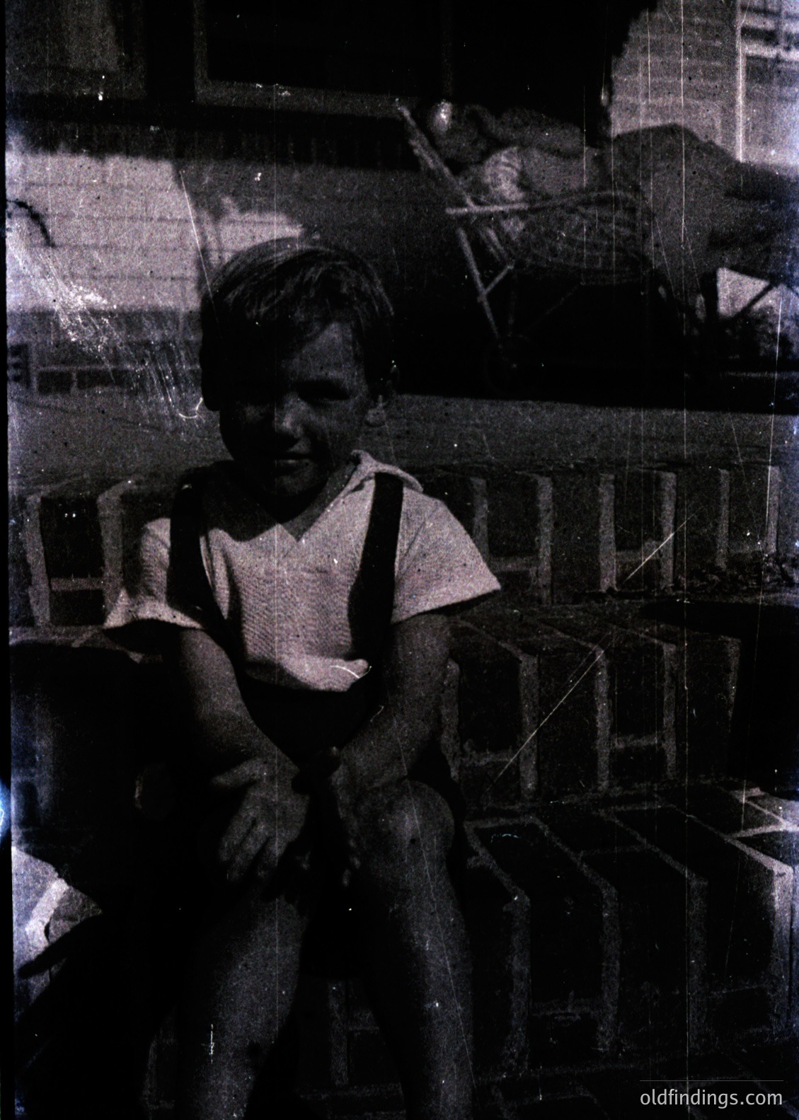 Vintage black-and-white photo of a young boy in suspenders, seated on a wooden chair indoors. Stacked crates and shelves with assorted items fill the background, suggesting a modest, utilitarian space. Likely mid-20th century (1940s–1960s), possibly Eastern European.