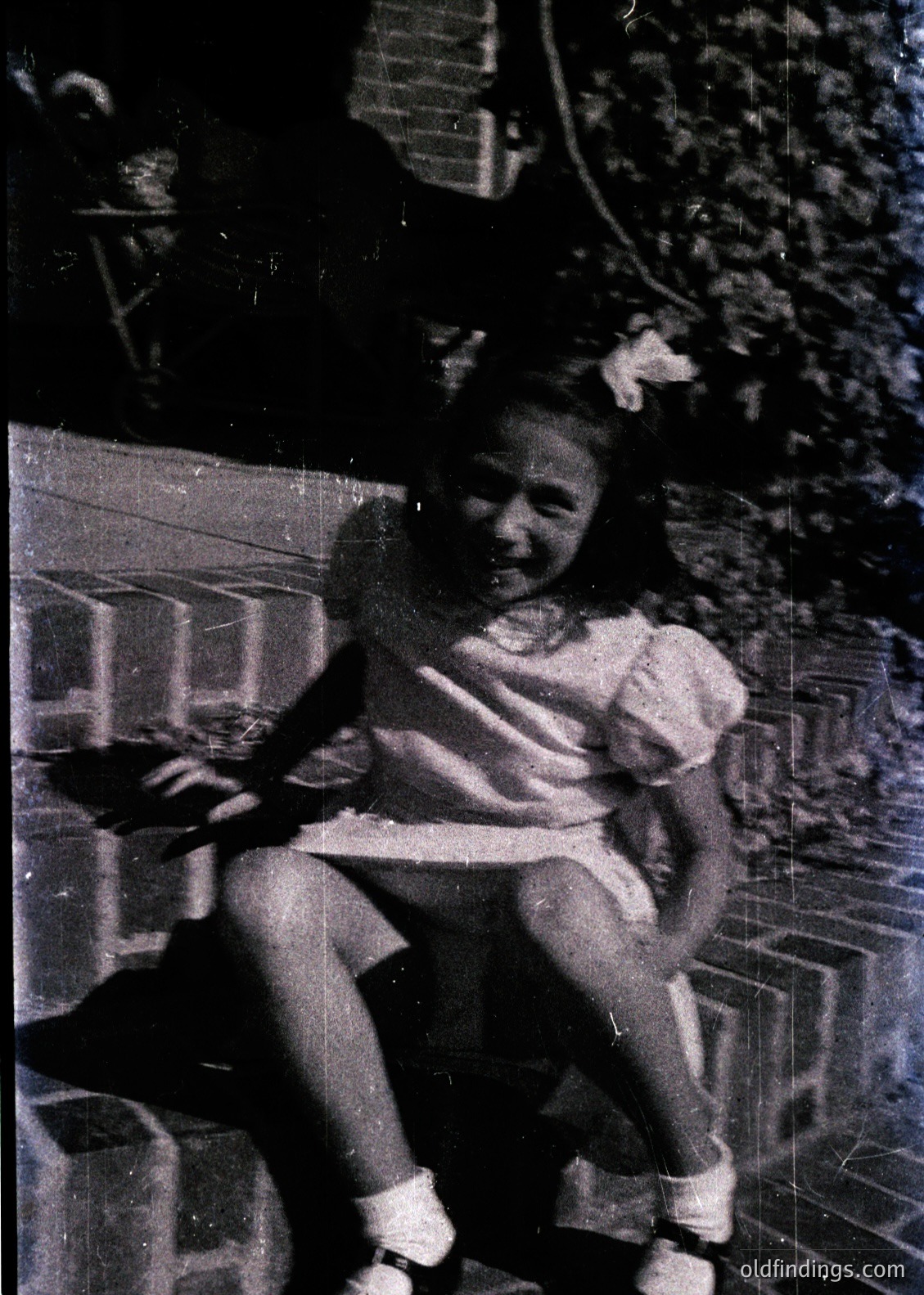Vintage black-and-white photo of a child in a 1950s-style dress with white socks and a headband, leaning on a tiled balcony railing. Indoor/outdoor setting with visible greenery and a window frame. Candid, nostalgic family portrait.