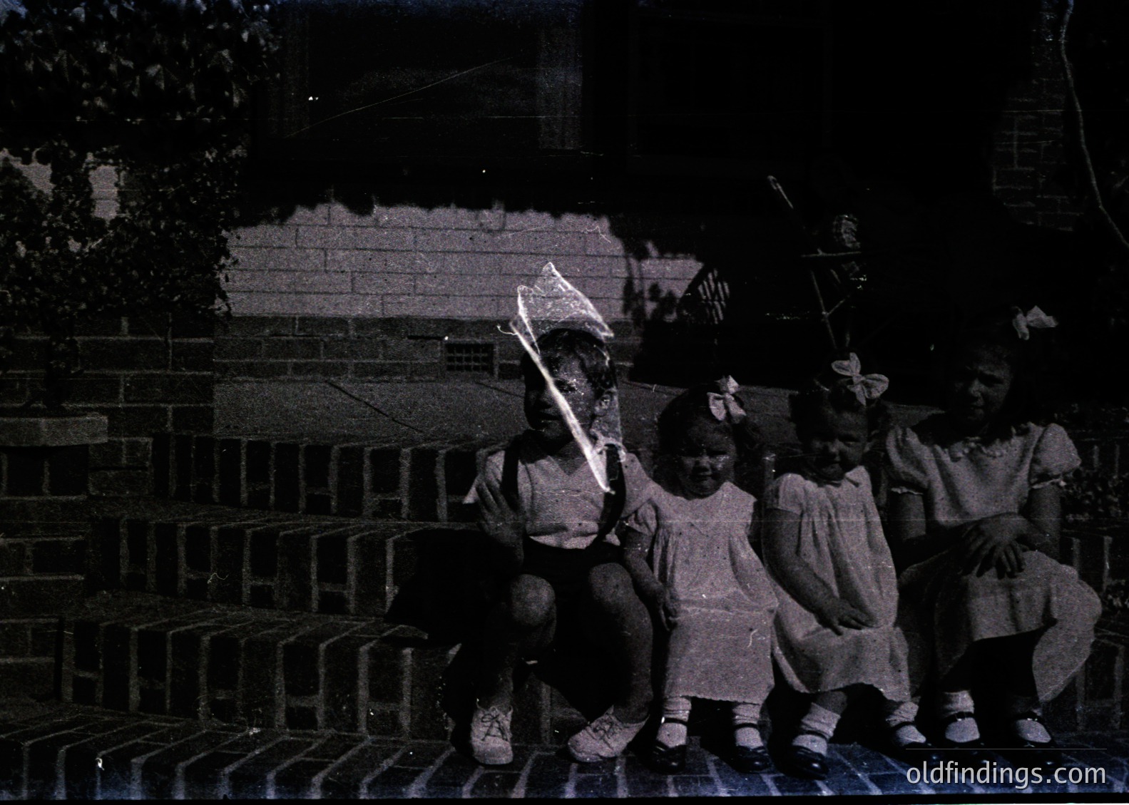 Four children pose on brick steps, holding kites in mid-19th to early 20th century attire—long dresses, bows, and cardigans. Urban setting with brick wall and greenery in background. Evokes nostalgia for early 20th-century childhood play.
