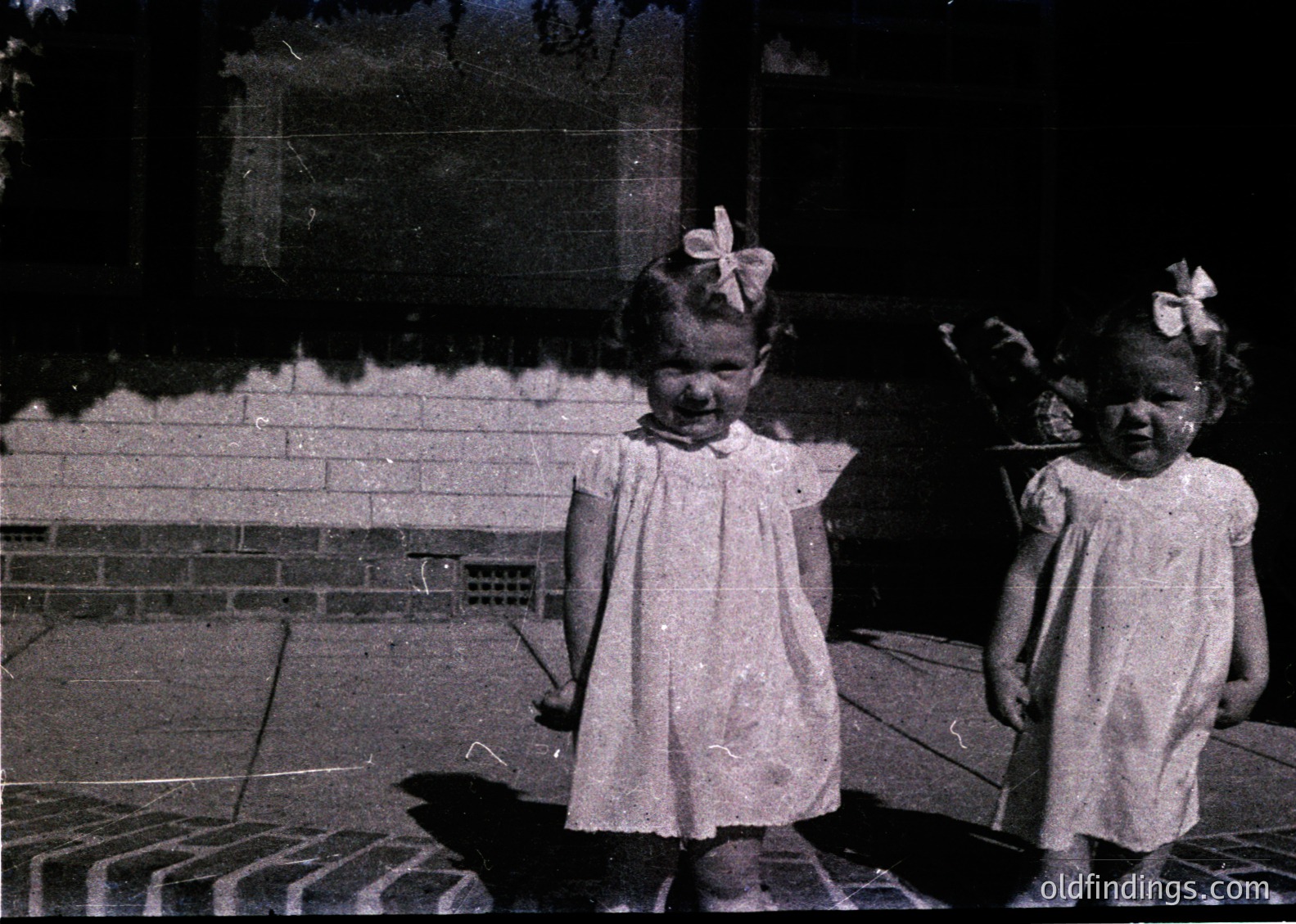 Two young girls in early 20th-century dresses with bows pose outdoors on concrete steps. Sepia-toned vintage photo suggests early photography era (pre-1920s). Simple dress styles reflect modest, practical fashion of the time.