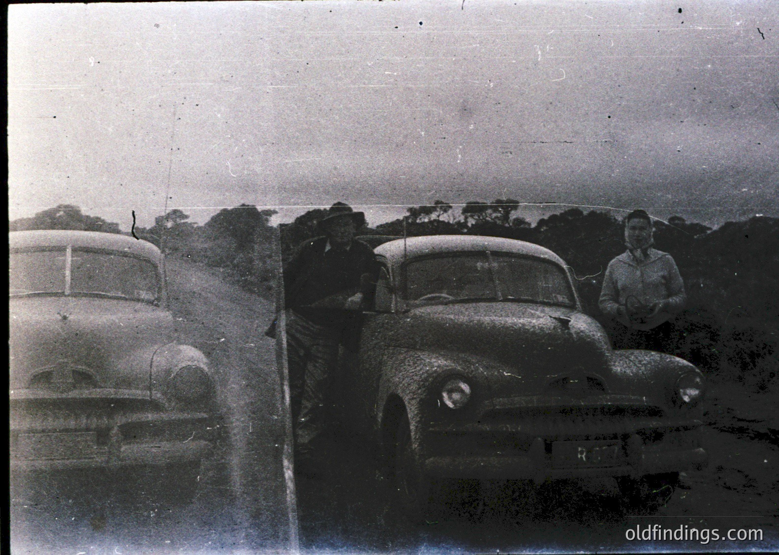 Vintage black-and-white photo of two classic cars parked on a rural road, likely 1950s. A man leans against the rear of a sedan with rounded fenders, while another stands beside a station wagon. Dense trees and overcast sky frame the scene, suggesting a forested area. Ideal for automotive history or nostalgic design references.