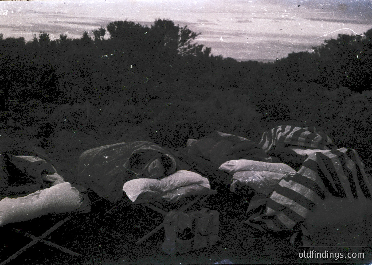 Vintage sepia-toned photograph of a battlefield scene with discarded military gear and equipment. Visible are helmets, canteens, and a partially visible uniform, likely from WWI or WWII. The terrain appears rugged with scattered rocks and vegetation.