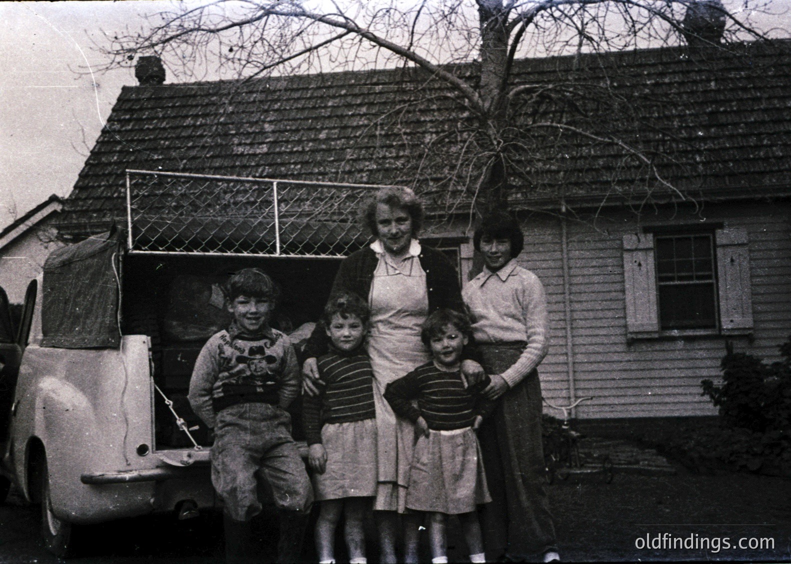 Family portrait in front of a mid-20th-century wooden house, likely 1950s–1960s. Five individuals pose near a vintage van, showcasing classic mid-century clothing (striped shirts, dresses, and overalls). The house features a gabled roof and simple wooden siding.