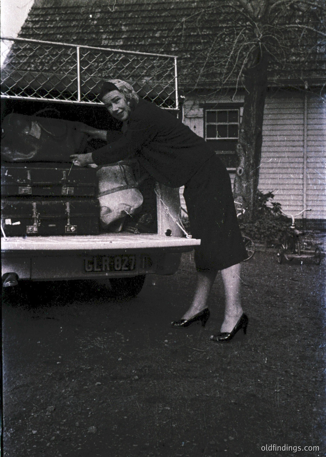 Mid-20th century woman in 1950s-style dress and high heels unloads luggage from a vintage truck with German license plate. Residential setting with brick driveway and wooden house. Classic post-war travel or relocation scene.