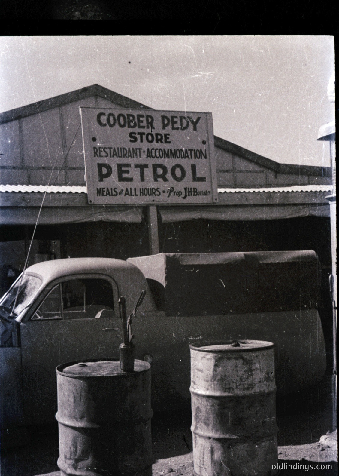 Vintage Coober Pedy petrol store signboard advertising meals, accommodation, and petrol, mid-20th century. Classic 1950s-60s outback truck parked beside rusted oil drums. Rustic Australian outback setting.