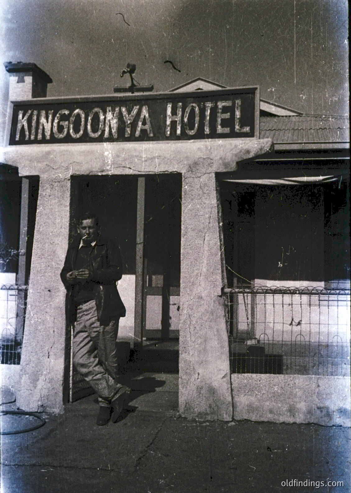 Black-and-white photo of a man in 1960s-era attire posing under the "Kingoonya Hotel" sign. Concrete columns frame the entrance, with a small balcony and barred window visible. Likely Australian due to the name.
