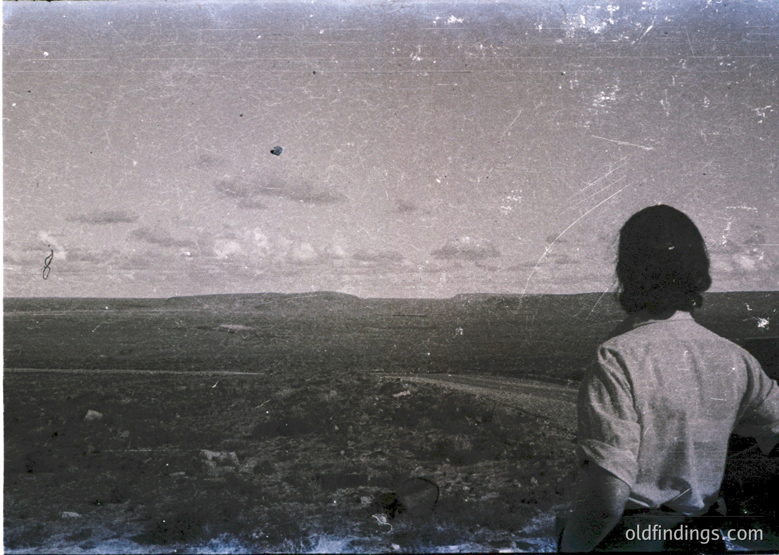 Vintage black-and-white photo of a lone figure seated on a cliff edge overlooking a rugged, barren landscape with sparse vegetation and distant hills. The person’s back faces the camera, wearing a loose, high-neck shirt. Damaged print with visible scratches and fading. Likely 1950s–1970s, possibly European or North American.