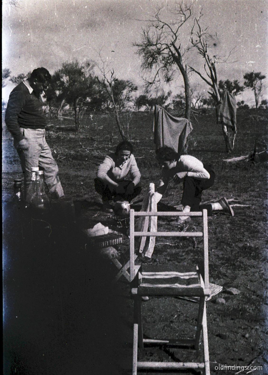Vintage black-and-white photo of three men in outdoor setting, likely 1940s-1950s. One man stands near a tree, another kneels beside a makeshift wooden frame with fabric draped over it, while a third sits on the ground. Clothing suggests utilitarian, possibly military or laborer styles. Background shows sparse trees and makeshift tents or tarps. Ideal for historical research on post-war reconstruction or textile work.