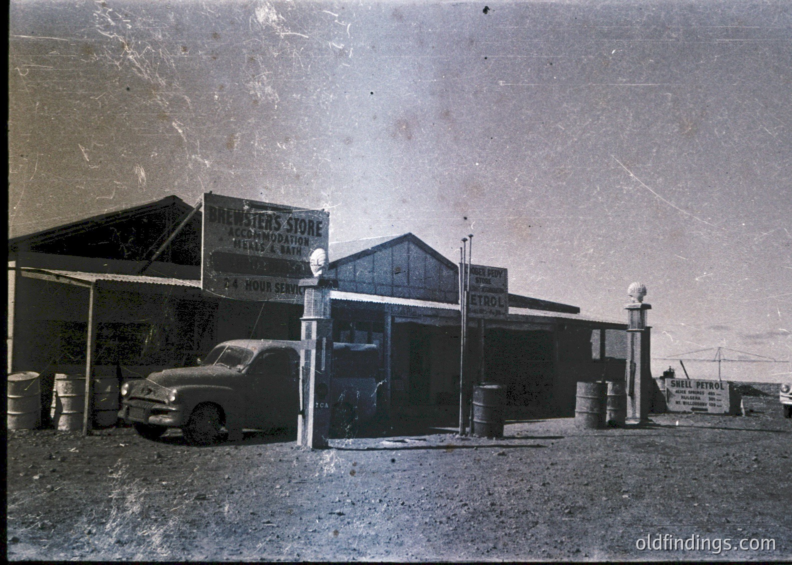 Vintage 24-hour service station with "Newman's Store" sign advertising auto repairs, tires & batteries. Classic mid-century truck parked beside fuel pumps and barrels. Rustic, rural roadside scene, likely 1950s–1960s USA.
