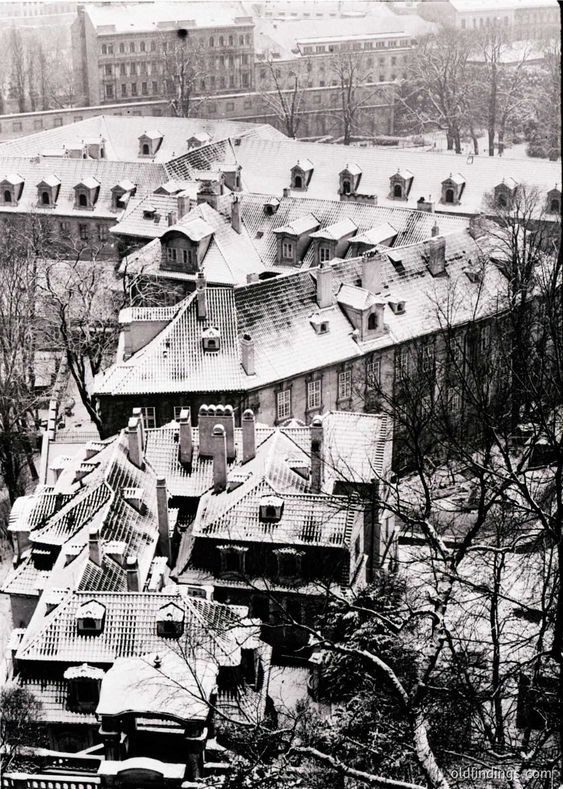 Snow-covered rooftops of a densely packed European cityscape, likely 1950s–1970s. Distinctive gabled architecture with chimneys and dormer windows. Leafless trees frame lower edge, hinting at winter season. Mid-rise institutional building visible in background.